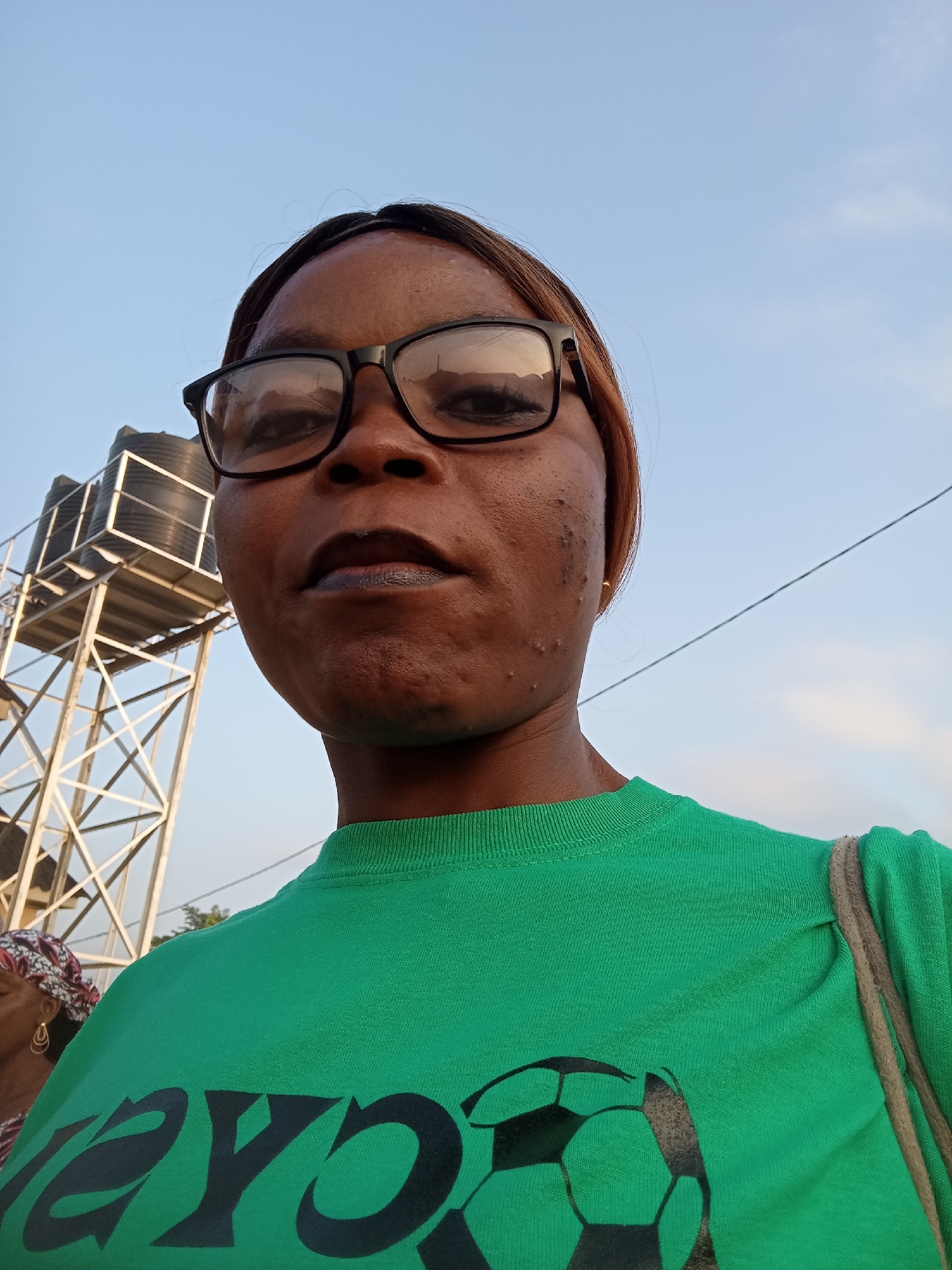 A waist-up, medium shot captures a woman of African descent from the chest up, facing the camera. She has dark skin, short, dark hair, and is wearing large, black-rimmed glasses. Her expression is neutral. She is wearing a bright green t-shirt with a black soccer ball design on the front. A thin, rope-like strap is visible over her right shoulder. In the background, a metal water tower with two black tanks stands to her left. The sky is clear blue with a few faint clouds. A thin black wire runs diagonally across the upper right portion of the frame. The scene appears to be outdoors, likely in a residential or industrial area, given the presence of the water tower. The image quality is fair, with good lighting and color saturation.