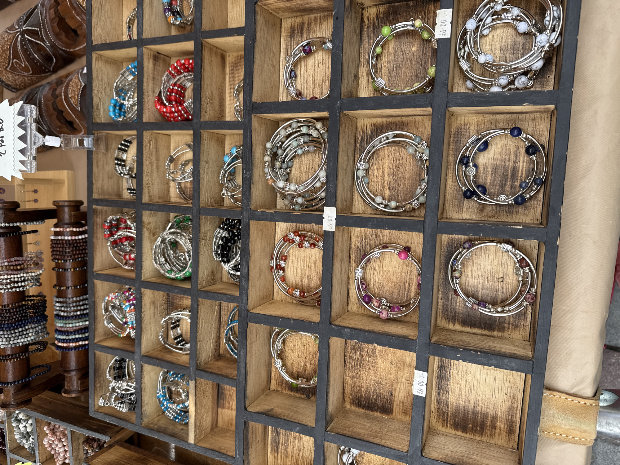 The image shows a display of beaded bracelets in a multi-compartment wooden tray at a market stall. The bracelets are of various colors and bead types, including blue, red, green, purple, and multi-colored beads. Some bracelets are silver-toned, while others appear to have different metal accents. Small price tags are visible on some of the compartments. In the background, more bracelets are displayed on wooden racks, along with some leather items that appear to be cowboy boots or similar footwear. The overall scene suggests a retail environment, likely an outdoor market. The location is identified as Darlington, United Kingdom, at the Darlington Hippodrome. The photograph was taken on August 19, 2023. There are no people visible in the image, and no actions or interactions are taking place. The environment is a market stall, with goods displayed for sale.