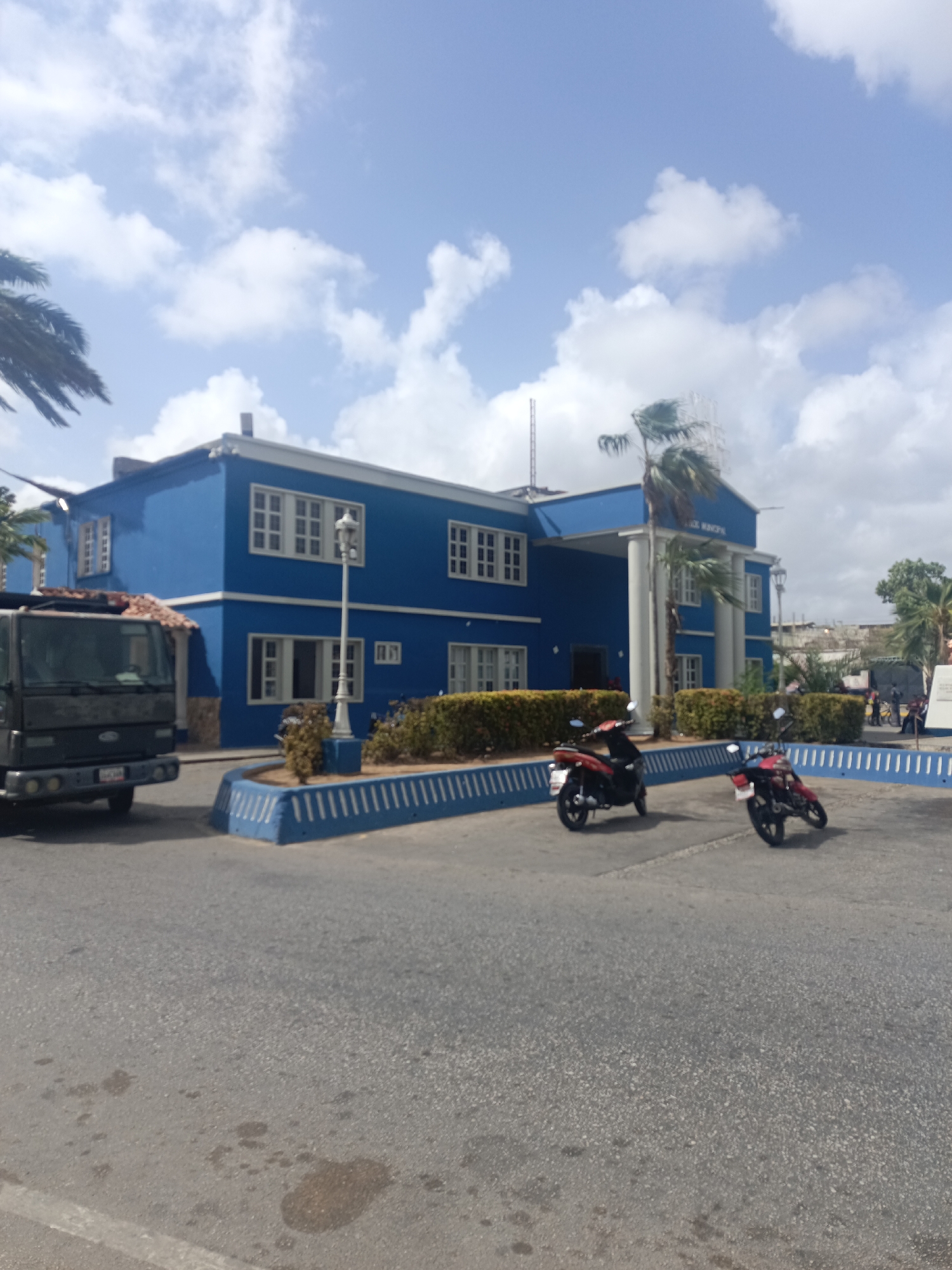 The image depicts the Alcaldía Municipal de Marcano (Marcano Municipal Hall) in Juan Griego, Venezuela. The building is two stories tall, painted bright blue with white trim, and features white columns on its facade. A large, dark-colored truck is parked on the left side of the frame, partially obscuring the side of the building. Two red motorbikes are parked in front of the building. A curb and a row of bushes border the building. The sky is partly cloudy. The pavement is visible in the foreground.