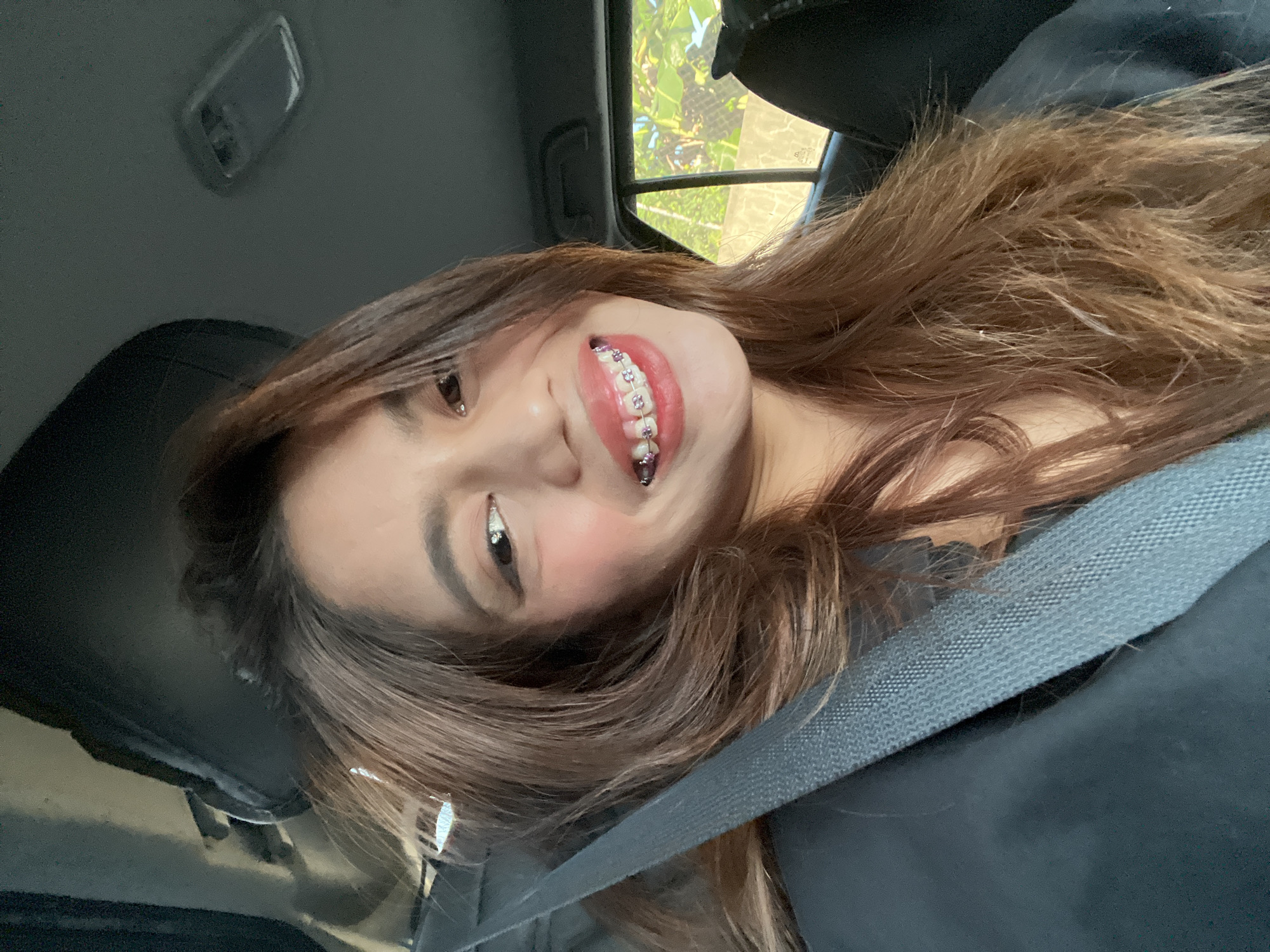 A young female, appearing to be in a vehicle, is captured in a close-up portrait, smiling directly at the viewer. She has long, wavy, light brown hair. Her upper teeth are fitted with clear orthodontic braces featuring purple elastic bands. She is wearing light makeup, including blush on her cheeks and what appears to be lip tint. A black seatbelt is visible diagonally across her left shoulder and chest, secured over a dark-colored garment. The car's interior ceiling is dark grey, with an overhead light fixture visible in the upper right quadrant of the image. Through the rear right window, lush green foliage and a light-colored concrete wall are discernible under bright daylight conditions. While the general environment is consistent with the Philippines, there are no specific visual identifiers within the image to confirm the precise location as Davao City.