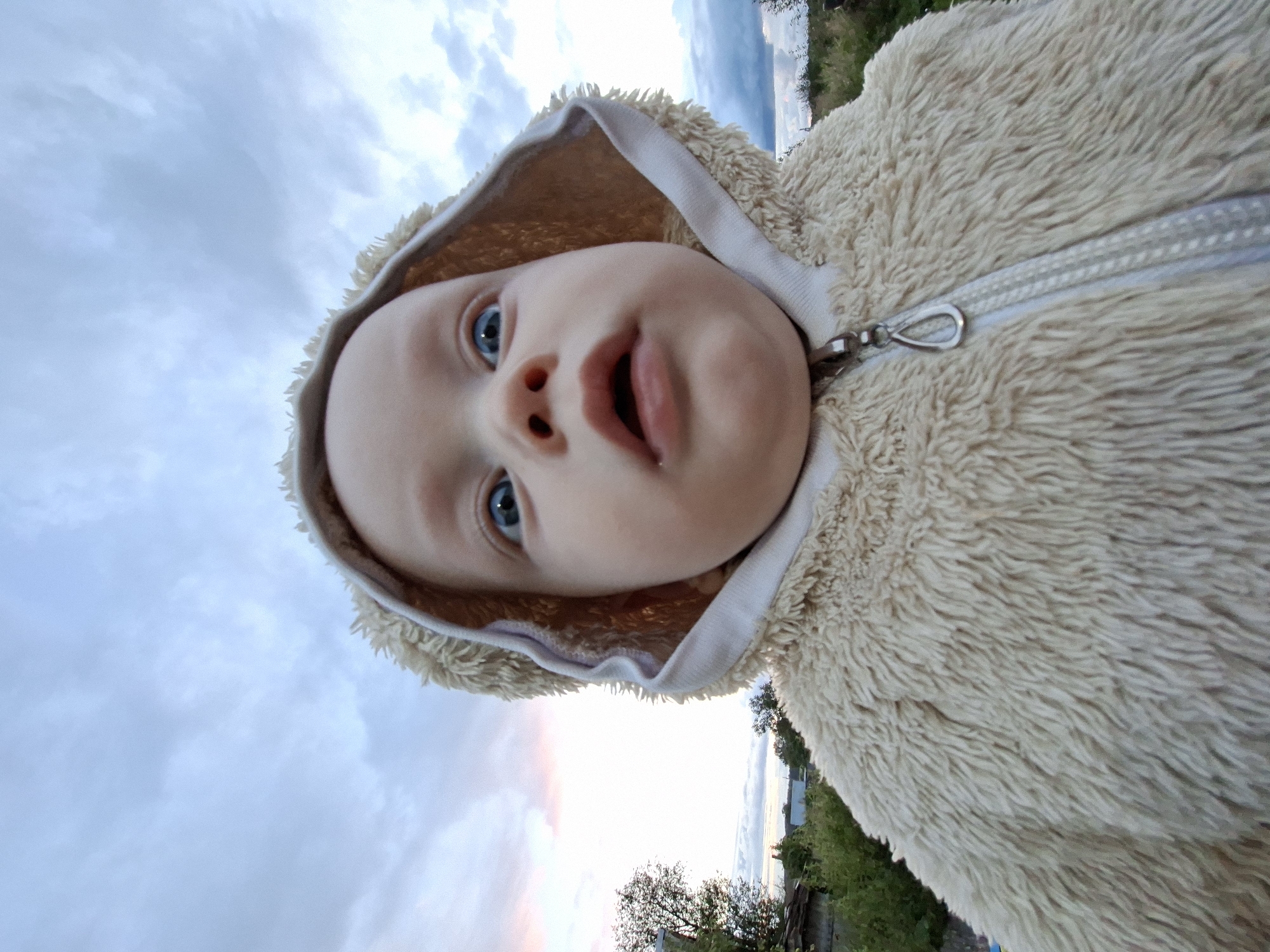 A baby is seen from a low-angle, close-up perspective outdoors, wearing a fluffy beige hooded jacket. The baby's face is directed towards the camera with wide blue eyes and an open mouth. The background consists of a cloudy sky with hints of pink and orange from a sunset, and a blurry view of greenery and structures. The image is taken in an outdoor setting that appears to be a rural or suburban area, possibly the city of Unknown, Unknown.
