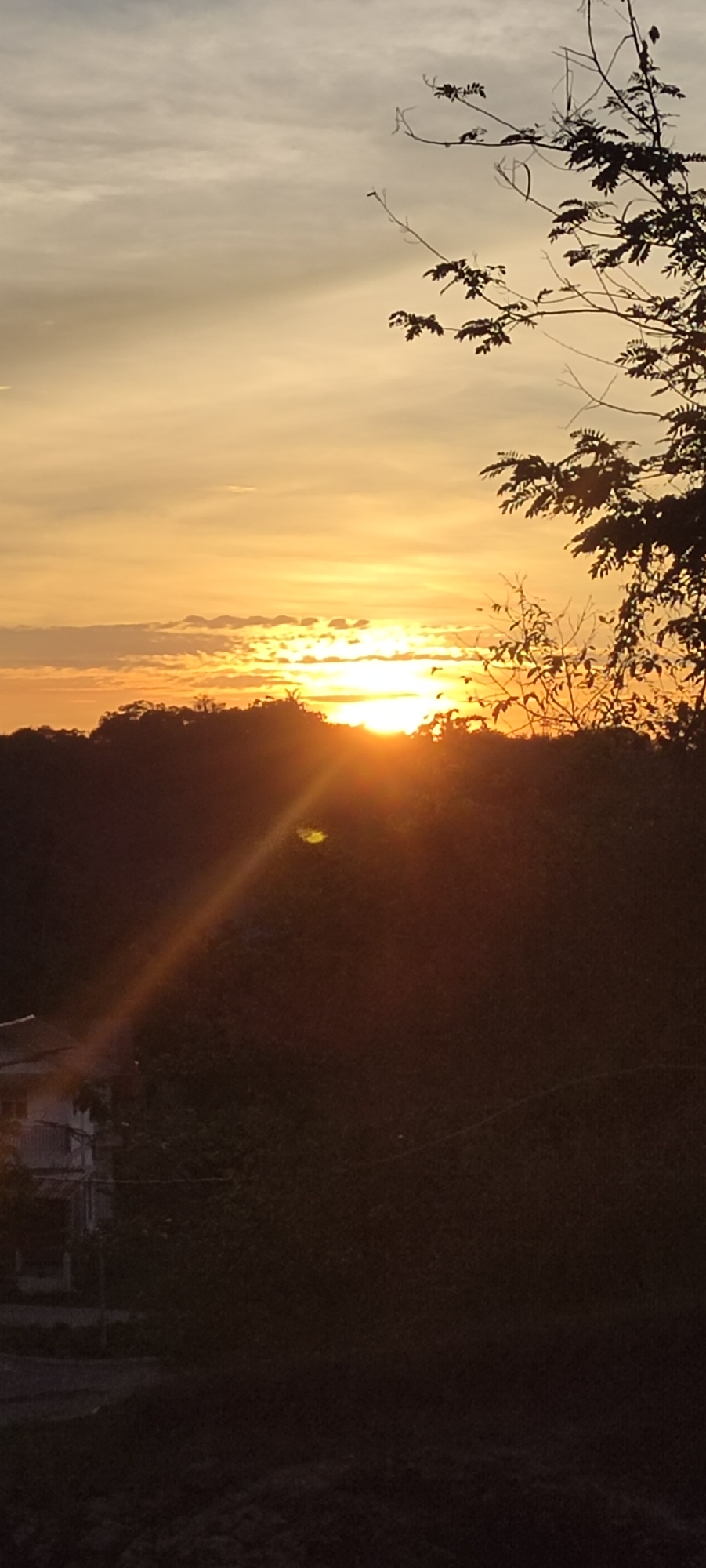 The image shows a sunset in Lhokseumawe, Indonesia. The sun is partially obscured by trees and vegetation. Light rays extend downwards towards a building to the left. The sky is a mix of orange and gray, with some clouds visible. Silhouetted branches are visible in the upper right. The foreground is dark, making the details difficult to discern.