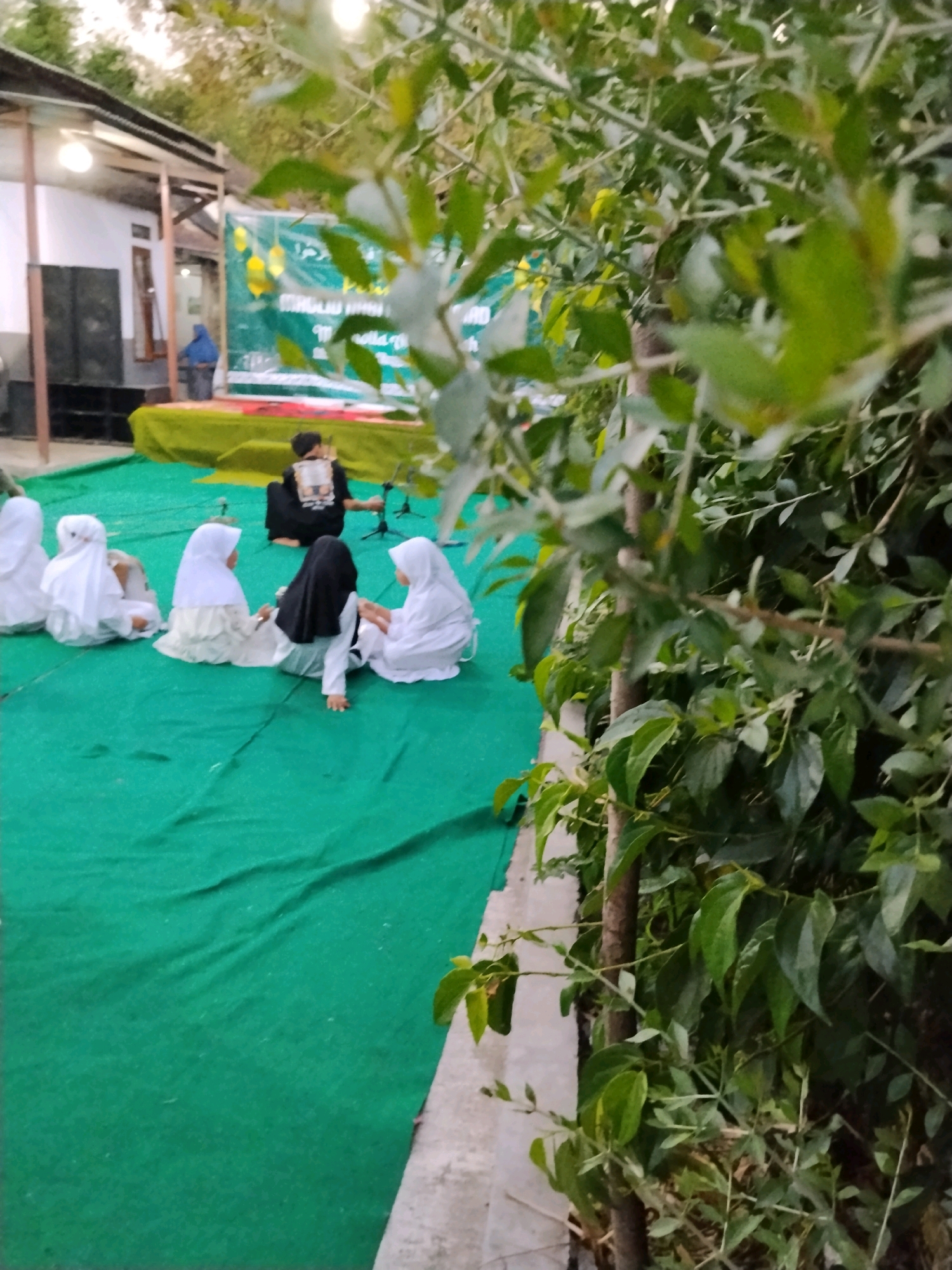 The image shows a group of young girls dressed in Islamic attire sitting on a green carpet. They appear to be observing an event on a raised platform in the background, where a person in black is seated and a banner with Arabic text is displayed. The event seems to be held outdoors in a setting that could be within the city of Kejawan, Indonesia, given the architecture and vegetation visible. The atmosphere suggests a cultural or religious gathering.