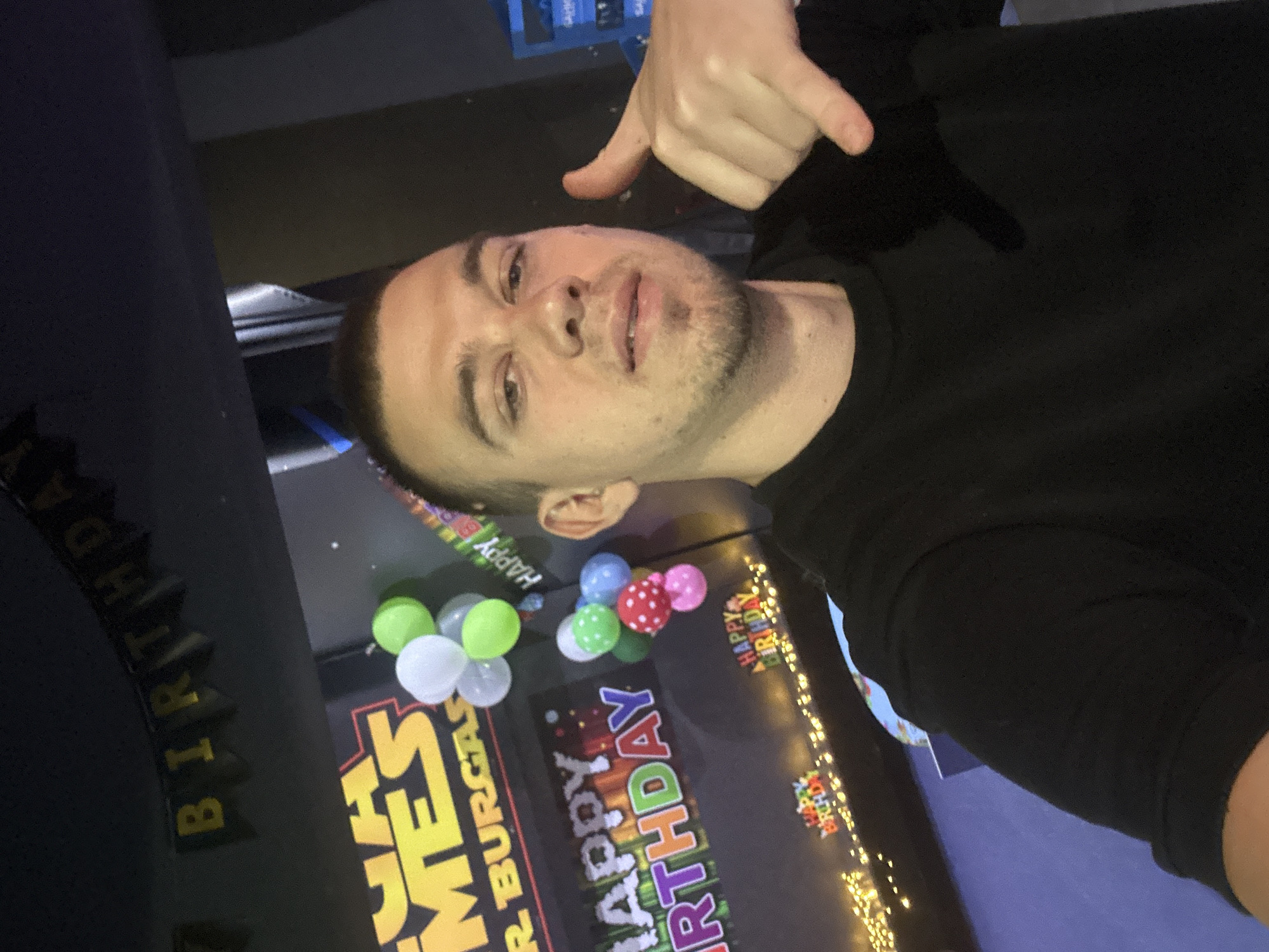 The image shows a young man taking a selfie indoors. He is wearing a black t-shirt and has short brown hair and a light beard. He is making a "shaka" hand gesture with his right hand. In the background, there are birthday decorations including balloons and banners that say "HAPPY BIRTHDAY". A banner with "MEGA GAMES" and "BURGAS" is also visible, suggesting the location is in Burgas, Bulgaria, possibly at an entertainment venue. The lighting is dim, typical of an indoor party setting.