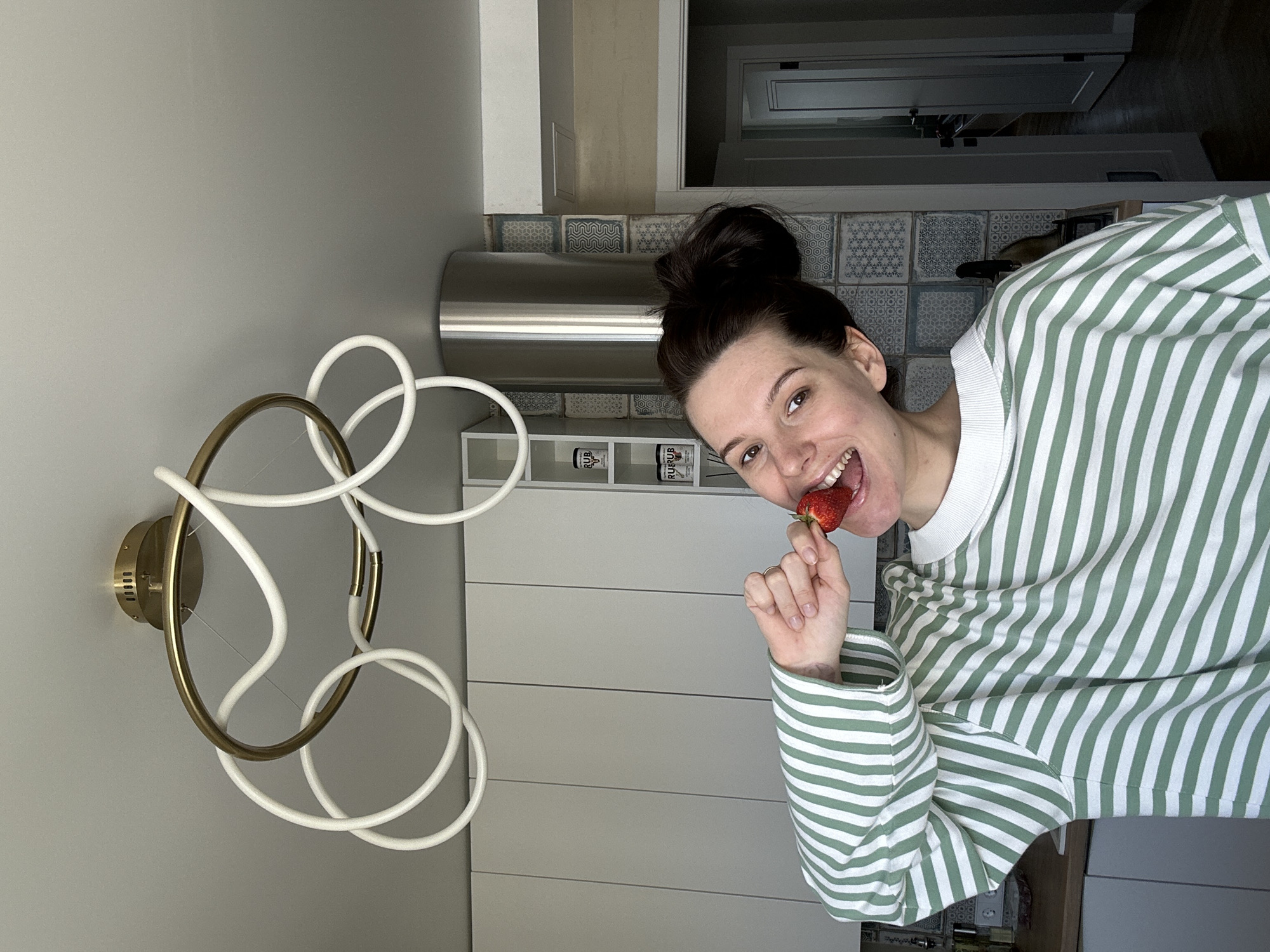 A woman is eating a strawberry in a kitchen. She is wearing a green and white striped shirt. The kitchen has gray cabinets and white appliances. There is a modern light fixture on the wall. There is tiling on the backsplash of the kitchen. There are also containers labeled "Rub Rub" on a shelf. The city of Chaikovsky, Russia is not explicitly mentioned in the image and cannot be confirmed.