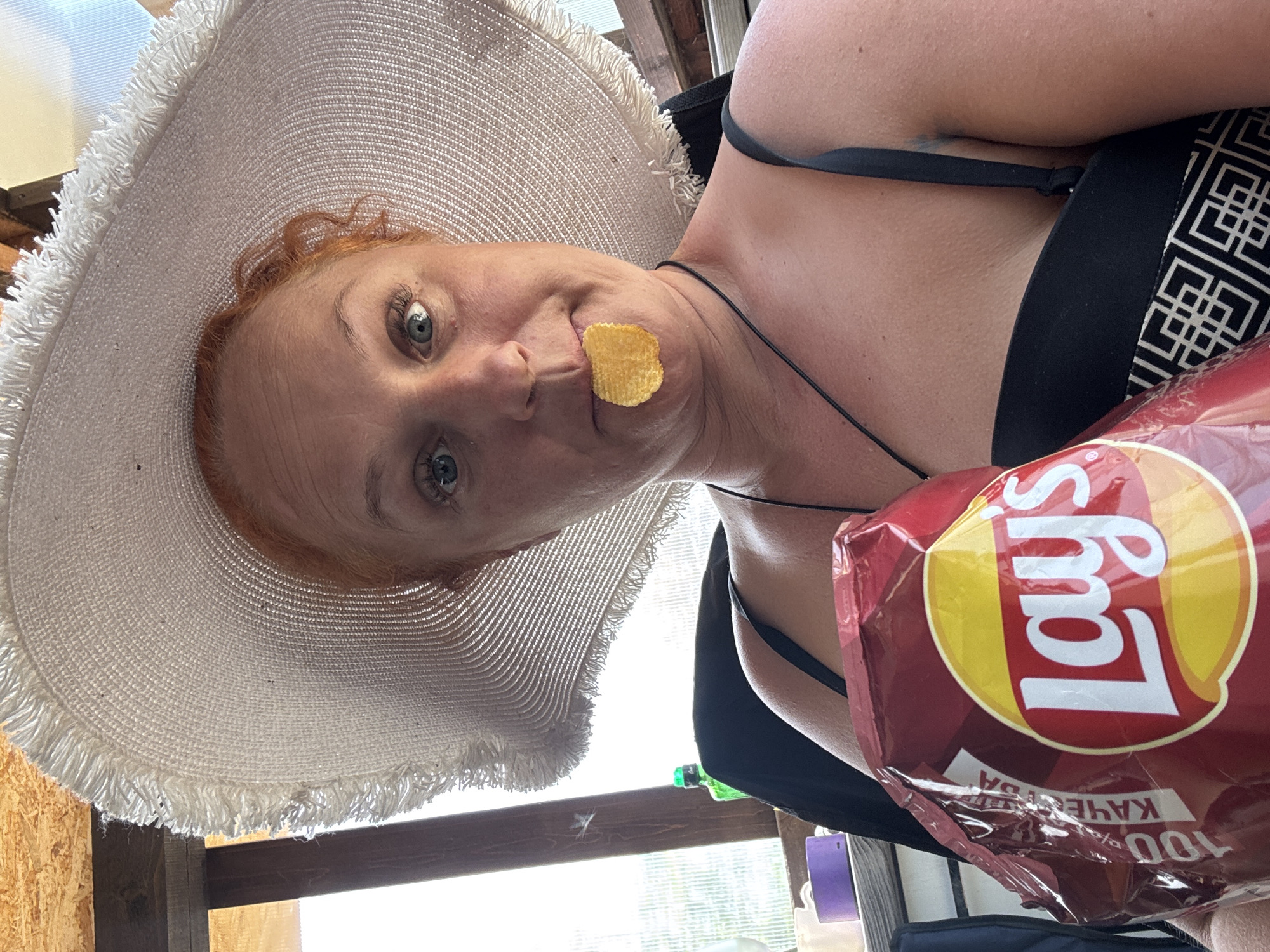 A middle-aged woman with red hair and blue eyes, wearing a large white sun hat and a black swimsuit top with a geometric pattern, is holding a bag of Lay's potato chips. She has a chip stuck to her face. The photograph appears to be taken from a low angle indoors, possibly at a sunny outdoor location. The bag of chips is written in russian with the description "100% картофель".
Since the bag of chips is written in russian and the photograph is potentially from a sunny outdoor location, it may be possible that the photograph was taken in Russia. However, this cannot be confirmed. It is not possible to confirm the location in the city of Chaikovsky, Russia.