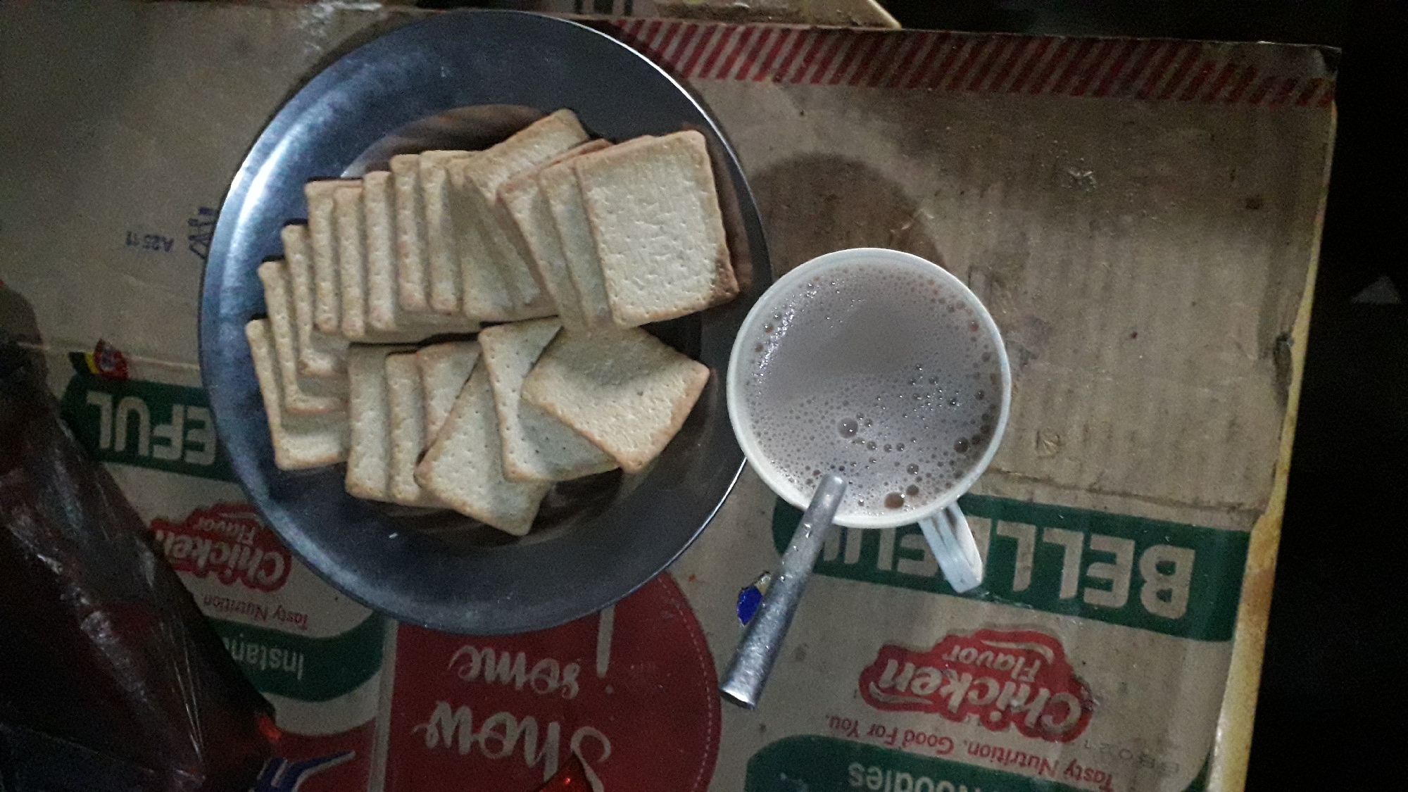 Here's a description of the image:
A top-down view shows a metal plate filled with stacks of square rusks. Next to the plate is a white mug filled with a frothy, brown beverage, possibly hot chocolate or coffee, with a metal spoon resting beside it. The entire scene is set on a cardboard box with partial text reading "BELLELL," "Chicken Flavor," "Tasty Nutrition," "Noodles," and "EFUL". The box appears to be a product packaging. The photograph appears to be taken in Onitsha, Nigeria.