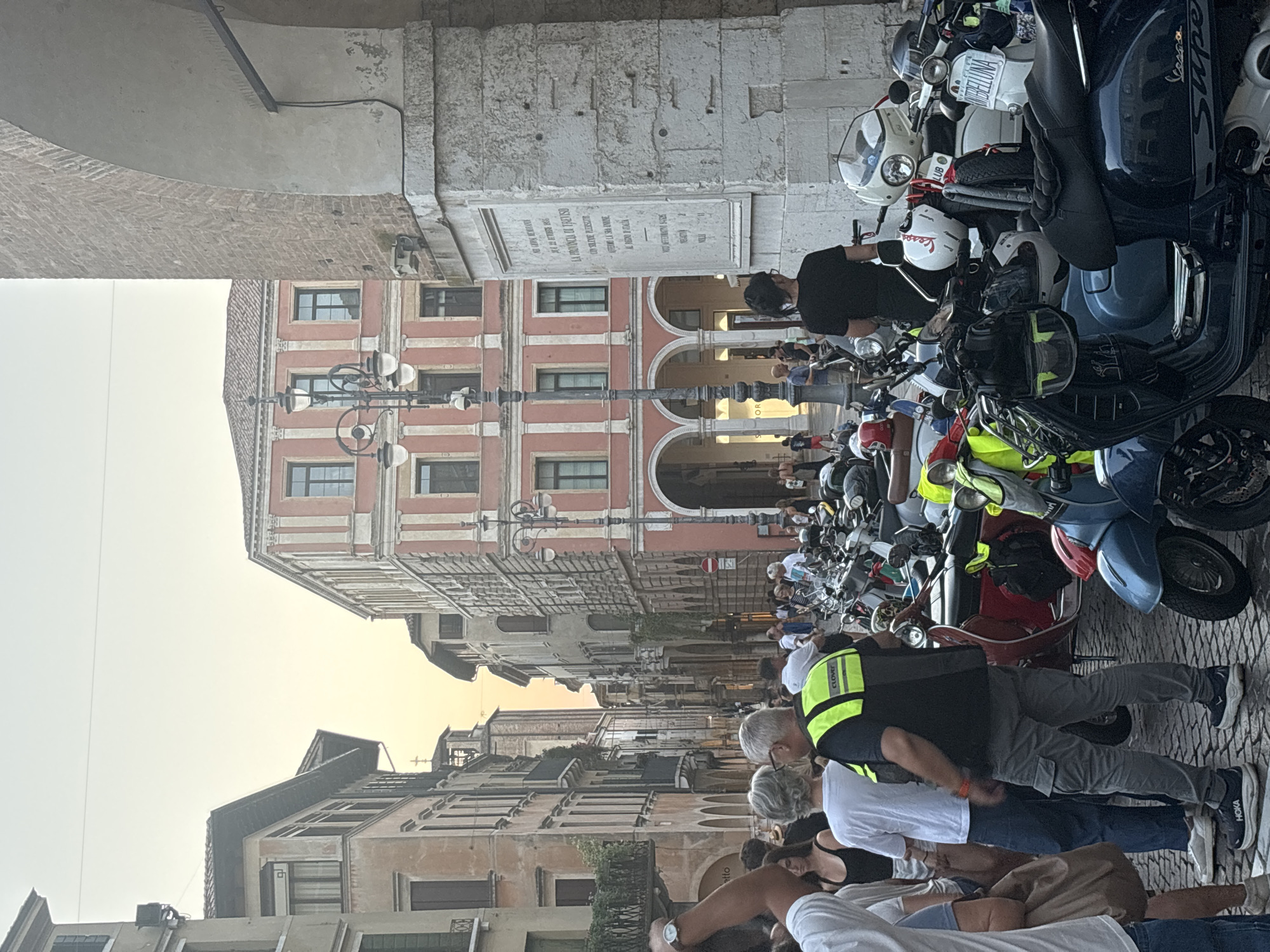 The image depicts a street scene in Treviso, Italy, with a prominent arcade on the right and buildings lining a narrow street. A cluster of Vespas and scooters are parked on the cobblestone street. Several people are visible, some standing and observing, others walking. A man in a bright yellow vest, likely a security guard or attendant, is standing in the foreground. The architecture suggests a historic European city, consistent with Treviso's character. The lighting indicates it is either early morning or late afternoon. The overall atmosphere is that of a bustling urban environment with a focus on local transportation and public spaces.