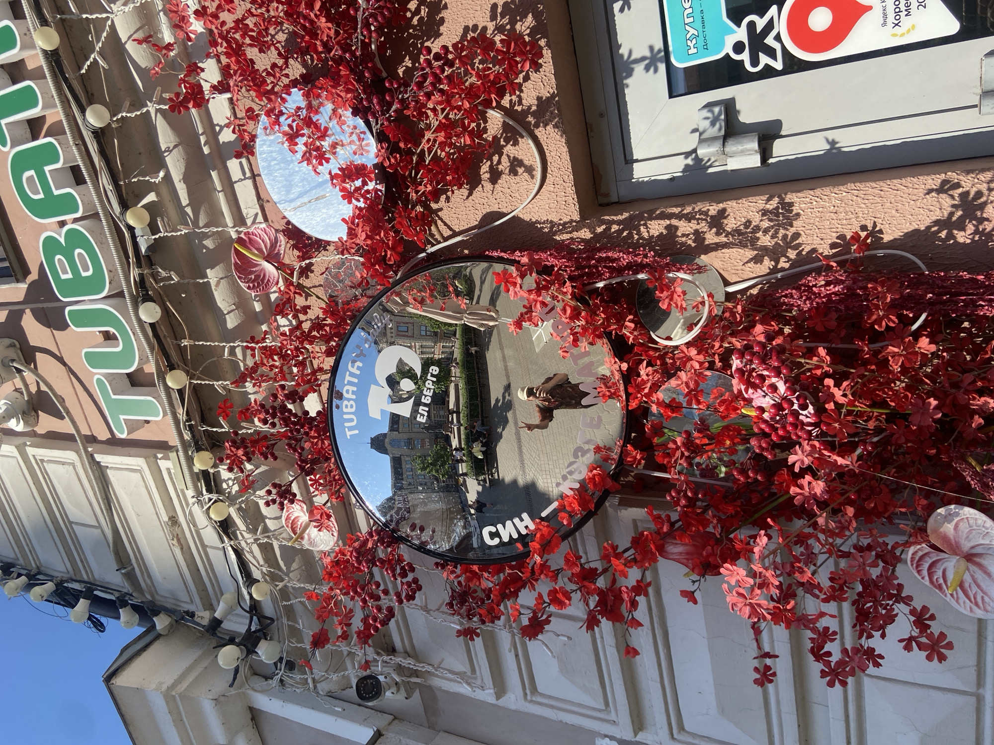 The image depicts an exterior wall in Kazan, Russia, decorated with artificial red flowers and reflective discs. A large circular mirror prominently reflects the street scene, including buildings and passersby. A person's reflection is visible in the mirror, holding up two fingers in a gesture. The words "TUBATA" and "El Berga SIN" are visible on the face of the mirror. To the left of the mirror, a sign in green letters is partially visible. A string of white lights hangs along the building's edge. The building has a pale pink exterior. A small white sticker is on the window above the arrangement.