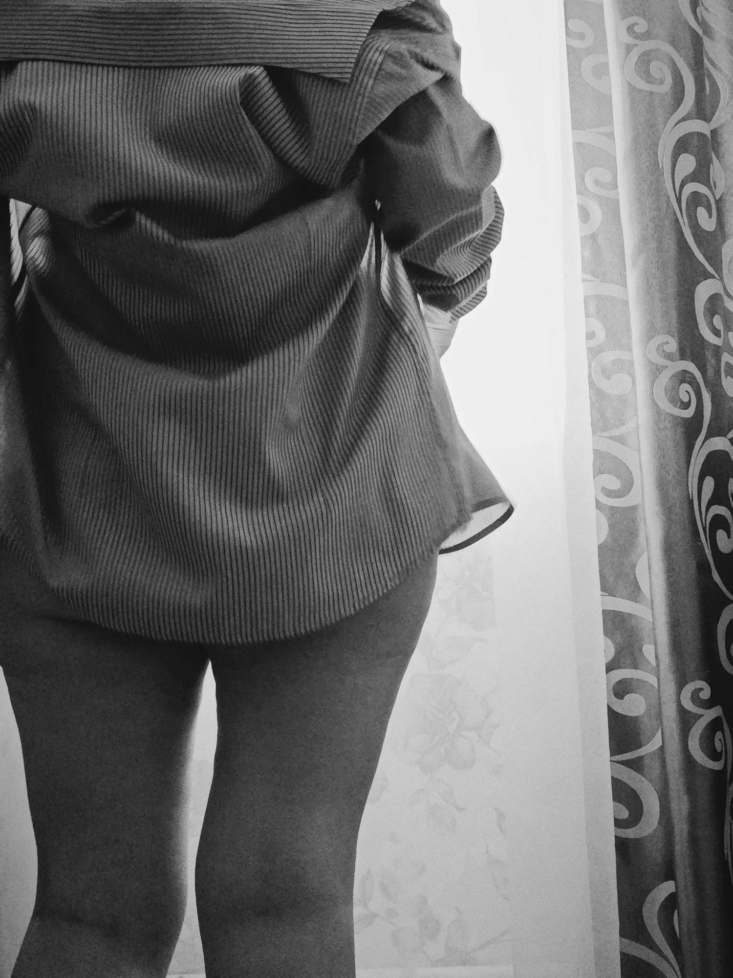 A black and white photograph captures an individual standing with their back to the viewer, positioned indoors in front of a window. The person is wearing an oversized, loose-fitting, long-sleeved collared shirt featuring a distinct vertical ribbed or striped pattern. The shirt's hem is above the mid-thigh, revealing the person's upper legs and thighs.
To the right, a window is partially visible, covered by two layers of curtains. The inner curtain is sheer and light-toned, with a subtle, barely discernible pattern. The outer curtain is darker and more prominent, adorned with a large, ornate swirling pattern. Bright light from the window illuminates the scene, creating contrast and outlining the person's silhouette on their right side. The setting appears to be a private room in Фоки, Russia. There is no visible text in the image.