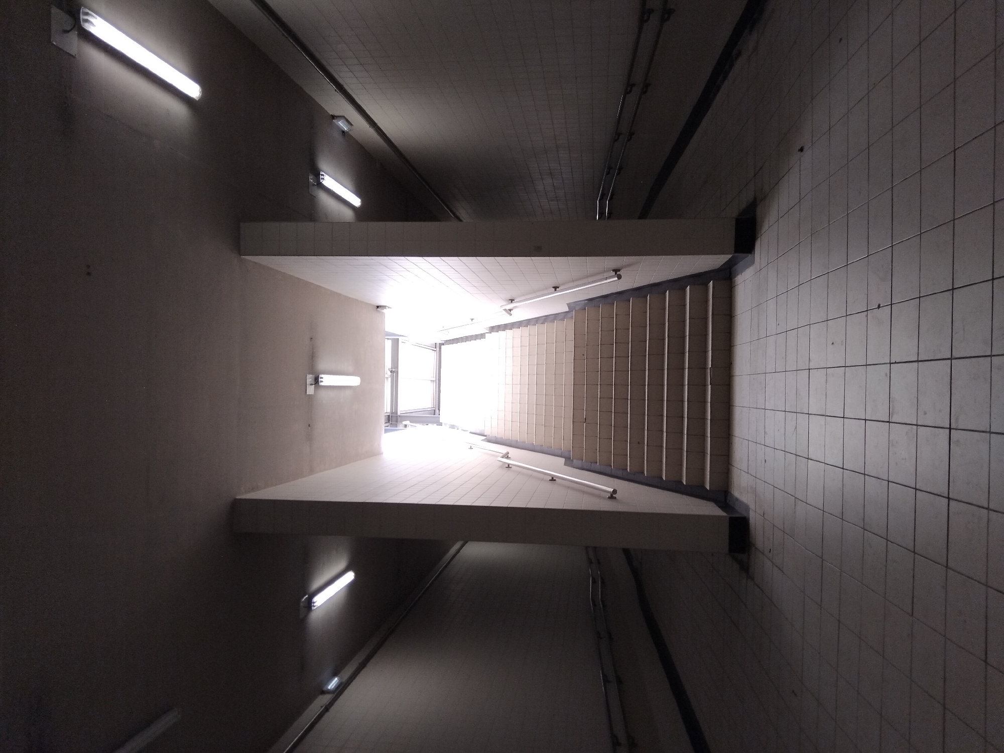 The image shows an underground pedestrian passage. The passage is tiled with light-colored square tiles on the floor and walls. The passage is illuminated by fluorescent lights on the ceiling. At the end of the passage, a wide set of stairs ascends towards natural light. On the left side of the passage, a dark, unlit corridor stretches into the distance, also tiled. On the right side, the passage is lit, and the stairs are visible. There are handrails on both sides of the stairs and along the walls of the dark corridor. The scene appears to be located in the city of Vila Nova de Famalicão, Portugal.