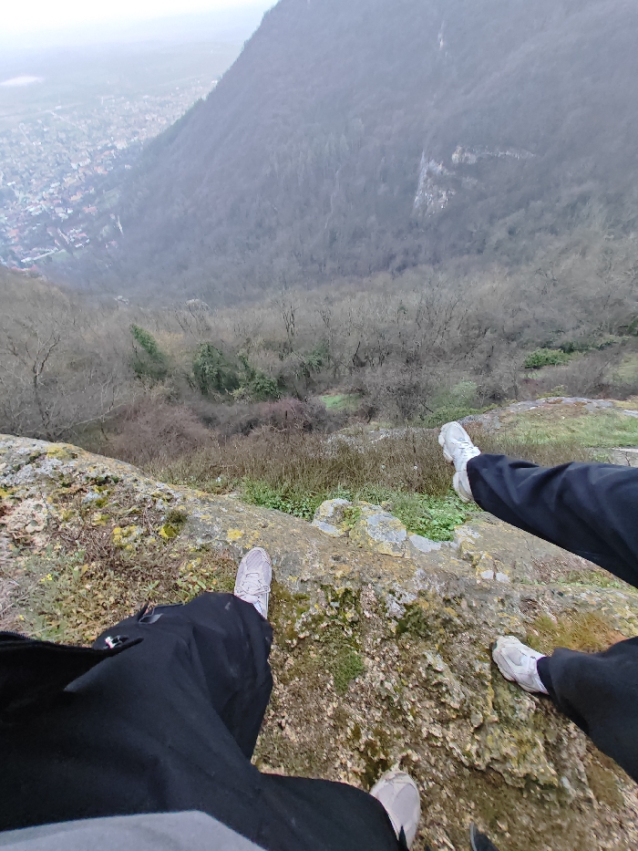 The image presents a high-angle view from a rocky precipice overlooking a densely forested valley and the city of Shumen, Bulgaria. In the immediate foreground, the lower bodies of two individuals are visible. One individual, likely the photographer, has their legs (wearing dark trousers and light grey/beige athletic shoes) positioned on a moss- and lichen-covered rock ledge at the very edge of a drop. To their right, another individual's legs, clad in dark trousers and white athletic shoes, extend horizontally outwards over the void. Below the cliff edge, a steep, tree-covered slope descends sharply into the valley. The trees are predominantly deciduous and appear bare, suggesting a winter or early spring season. In the distant background, nestled within the valley, lies the city of Shumen, characterized by a dense cluster of buildings with various rooftops, visible through a slight atmospheric haze. A large, tree-covered mountain forms the opposing side of the valley under an overcast sky.