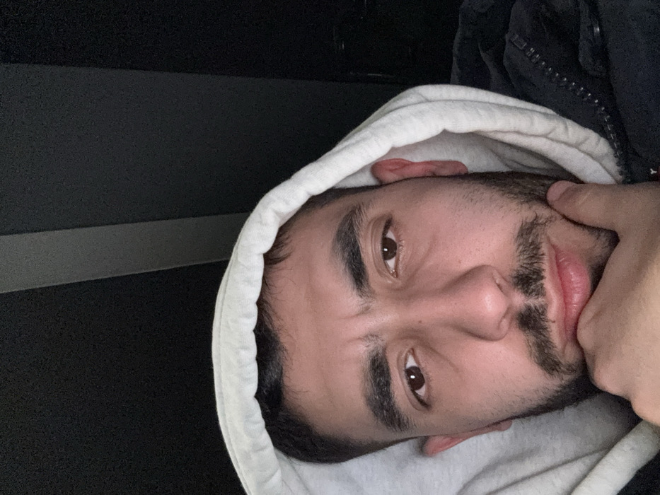 A close-up, eye-level photograph features a young adult male with dark hair, dark brown eyes, thick eyebrows, and a dark beard and mustache, looking directly forward with a neutral to slightly pensive expression. He wears a light grey or off-white hooded sweatshirt, with the hood pulled over his head, layered over a dark-colored zippered jacket, visible on his right shoulder and chest. His left hand is raised, with fingers resting against his chin and jawline. The background is uniformly dark, appearing to be an interior space or a low-light environment, with a single faint vertical grey line visible in the upper right quadrant. The specific location within Trieste, Italy, cannot be determined from the visual information provided.