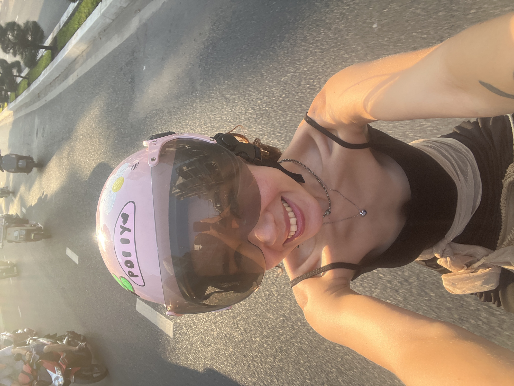 A woman with a pink helmet is riding a motorcycle on a street in Tay Nha Trang Ward, Vietnam. She is wearing a black top with thin straps and a necklace. Other motorcycles are on the road. The light suggests it is either morning or evening. There are trees along the roadside. The image appears to be a selfie taken while riding.