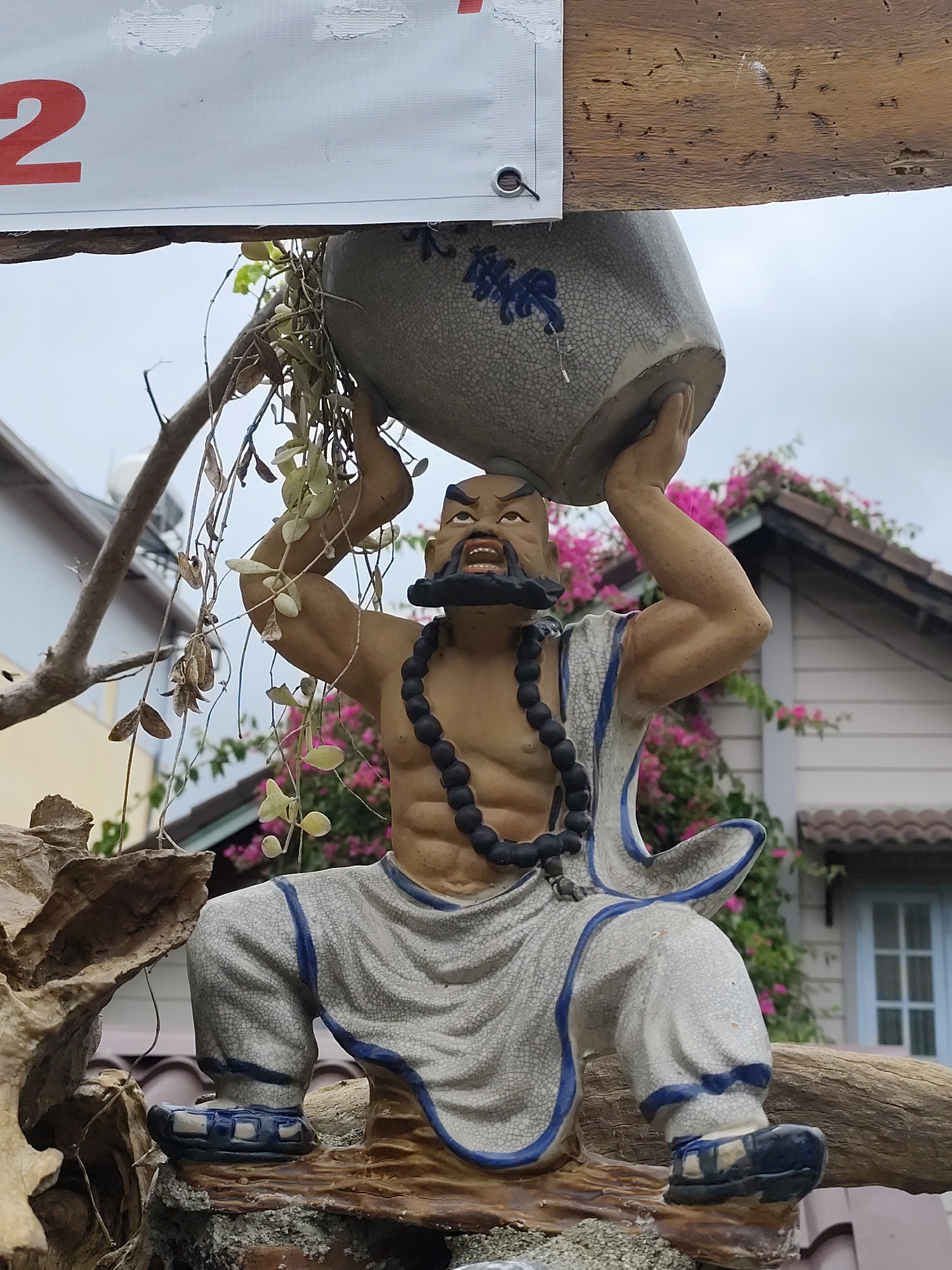 This image shows a ceramic statue of a man holding a large pot over his head. The statue is positioned outdoors, with buildings and lush pink flowers visible in the background, suggesting a residential or garden setting. The statue is weathered, with visible cracks on its surface, and is adorned with a dark beaded necklace and a white robe with blue trim. A potted plant with trailing vines hangs from a branch above the statue, partially obscuring the pot it holds. The scene is likely located in Da Lat, Vietnam, a city known for its temperate climate and abundant floral gardens, which aligns with the visual elements presented in the photograph. The overall impression is of an artistic display in a naturalistic or garden environment.