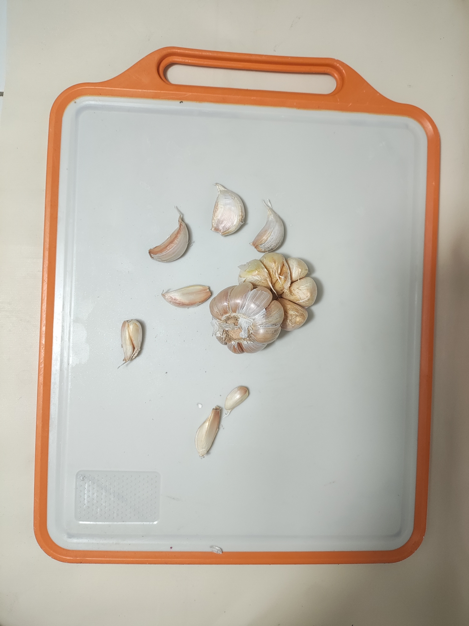 A still life photograph shows a cutting board with garlic on it. The cutting board is white with an orange rim and a built-in handle. It is placed on a light-colored surface. Scattered across the cutting board are several cloves of garlic, some still attached to a bulb. The garlic is light brown and beige. The image is shot from a top-down perspective. The location of the image in Wanajaya, Indonesia, is not discernible from the photograph itself.