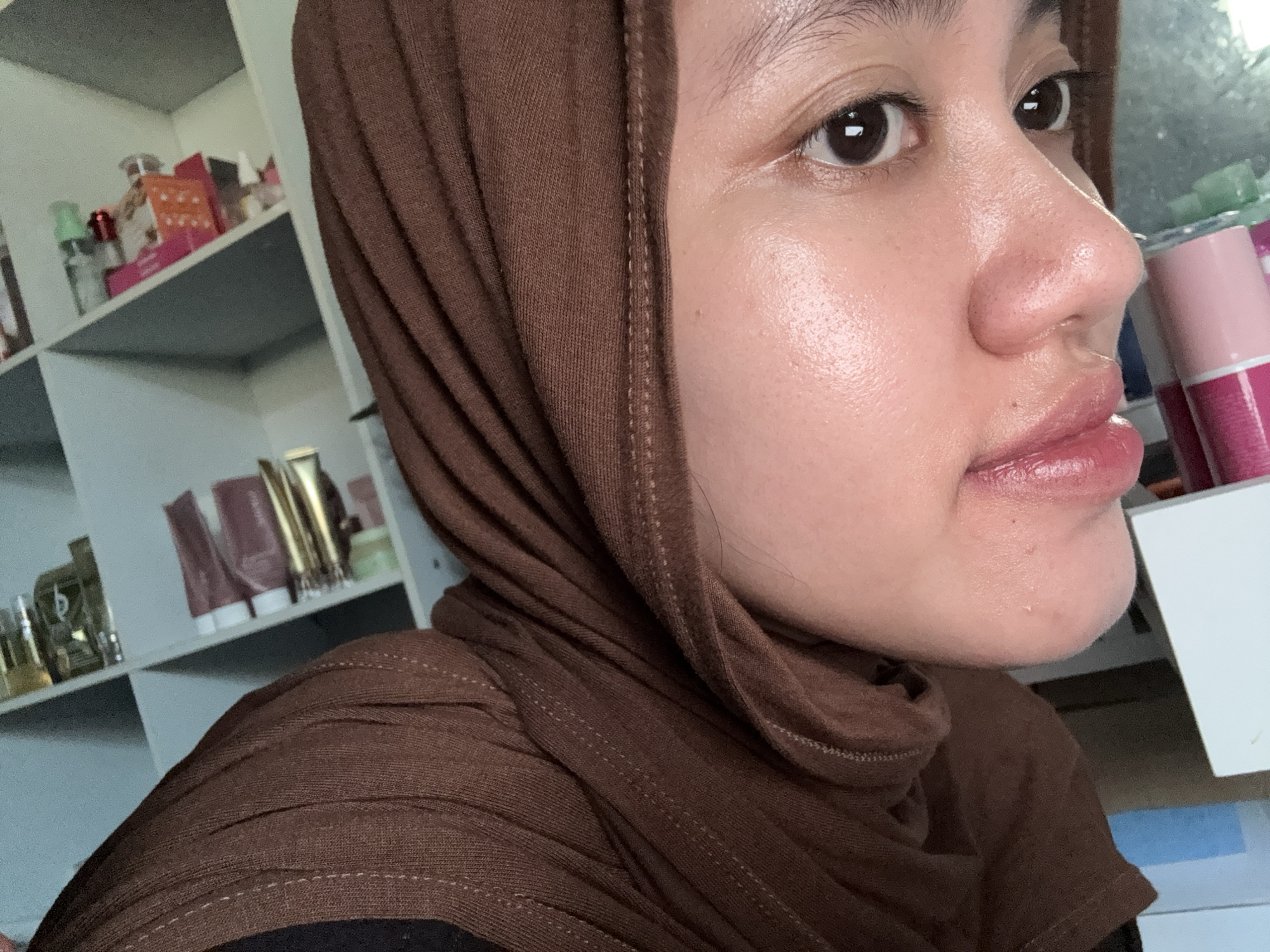 A close-up side view of a person wearing a brown headscarf. The person has brown eyes, a small mole on the left cheek, and glossy lips. Behind the person is a white shelf with various cosmetic products and containers on display. The environment suggests an indoor setting, possibly a home. There are no specific details to confirm the location in Gadingrejo, Indonesia.