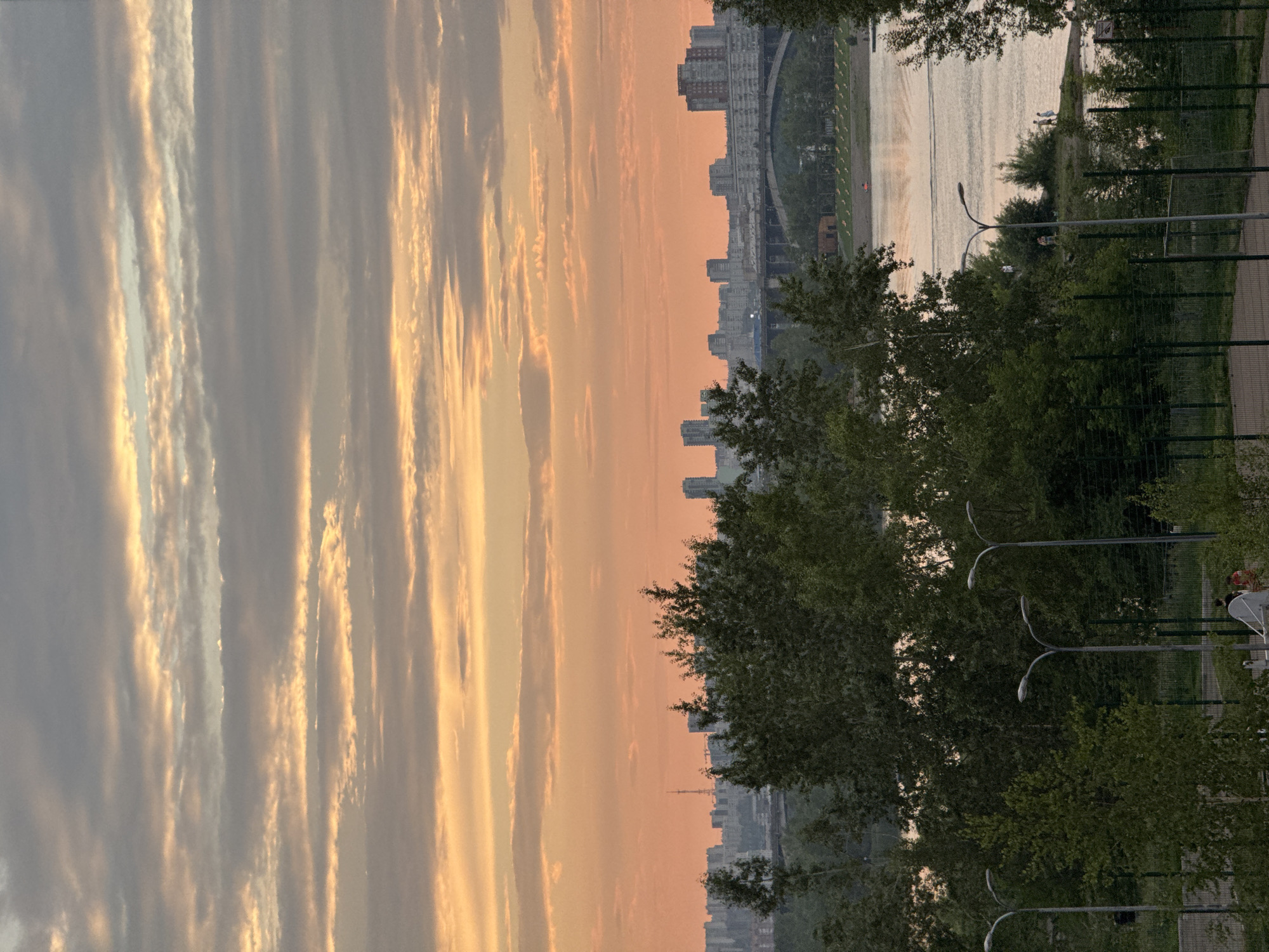 This image captures a serene sunset scene over a body of water, likely a river, with a city skyline in the background. The sky is painted with warm hues of orange and yellow, illuminated by the setting sun, with wispy clouds creating a dramatic effect. The cityscape features numerous apartment buildings and other structures, suggesting an urban environment.
In the foreground, lush green trees and foliage create a natural frame, contrasting with the man-made structures. A fence runs along the right side of the image, hinting at a park or recreational area. Several lampposts with distinctive curved arms are visible, indicating a public space.
On the bank of the water, a few individuals are present, suggesting a leisurely evening activity. The calm water reflects the soft light of the sunset, creating a peaceful atmosphere. The overall mood is tranquil and contemplative, with the soft lighting and natural elements contributing to a sense of calm. The time of day is clearly sunset, given the warm, low-angled light and the colors in the sky. The location cues, such as the cityscape and the presence of a river, along with the provided context of Krasnoyarsk, Russia, help to place the scene. There is no visible text in the image.