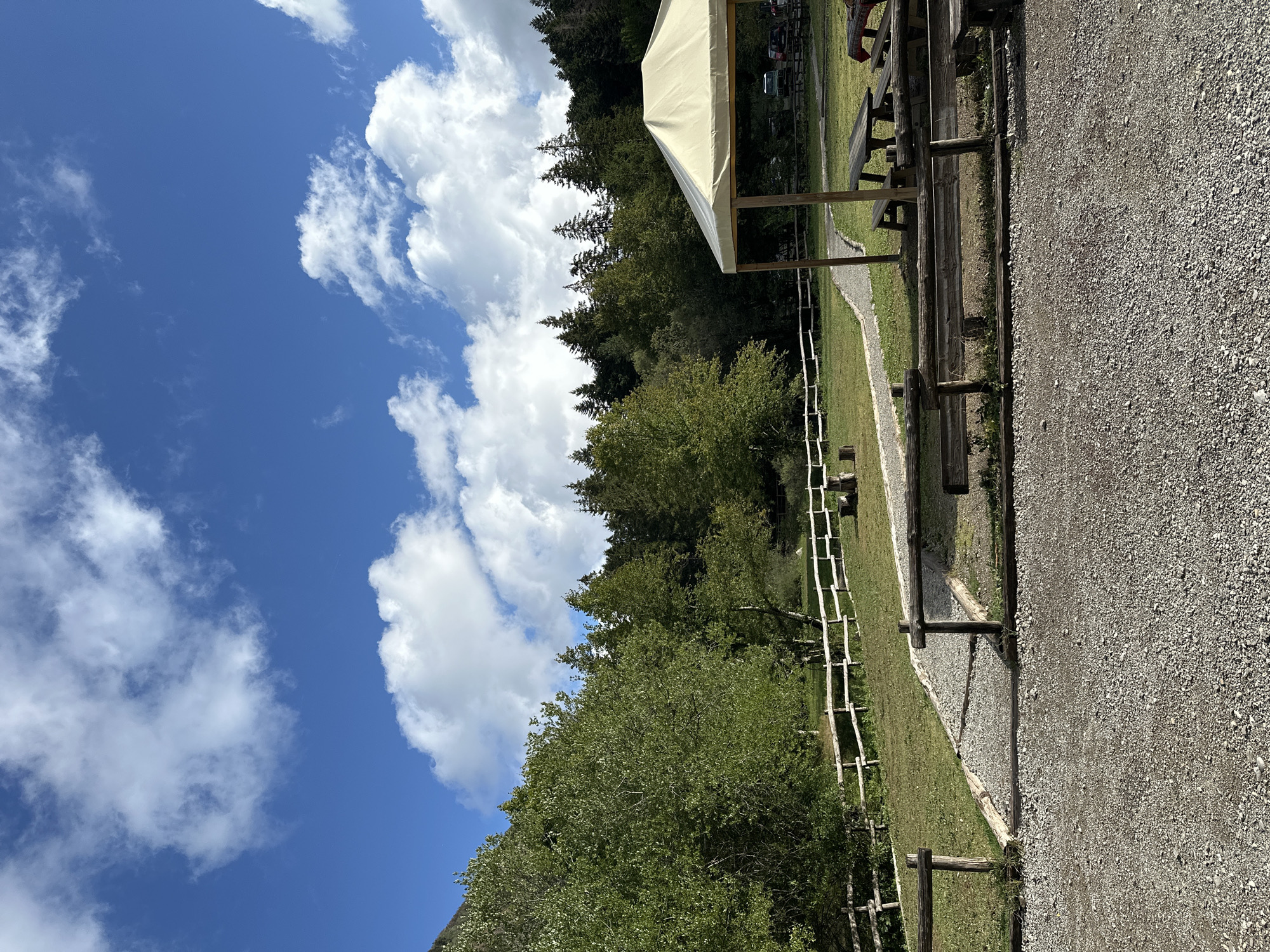 In Corniglio, Italy, a picnic area is depicted under a partly cloudy sky. The foreground is a gravel path, and beyond it, a wooden fence separates the path from a grassy area with a walking path. To the right, a white-tented picnic shelter covers several wooden picnic tables and benches. In the background, a dense forest of green trees borders the area, with a steep, wooded hillside rising behind it. The scene suggests a tranquil outdoor recreational space.