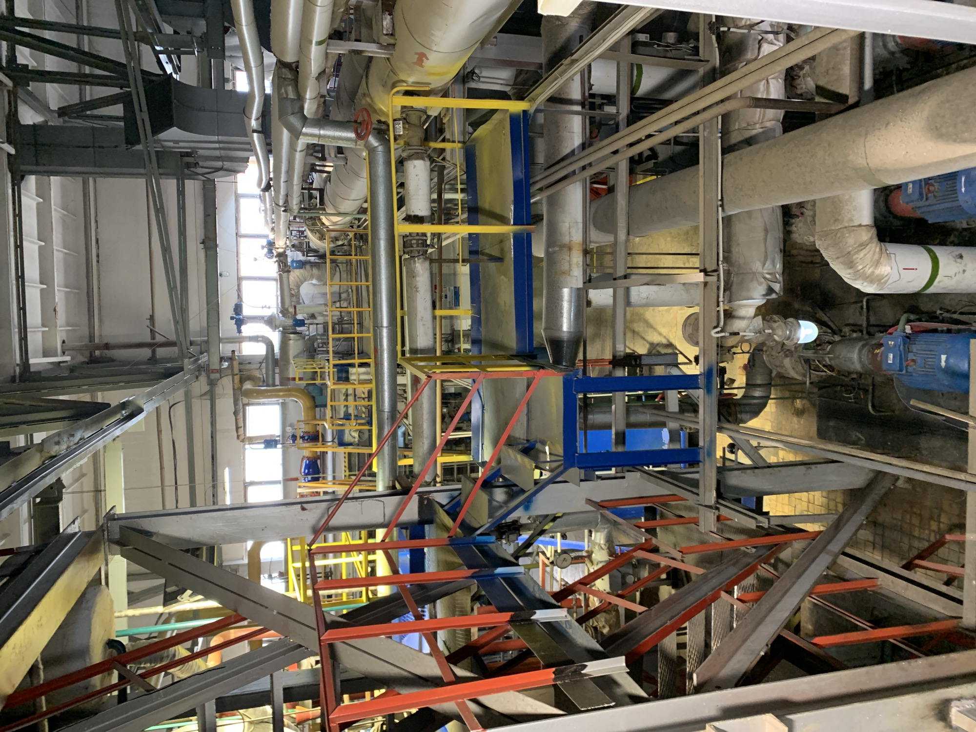 This image depicts the complex interior of an industrial facility, likely a power or chemical processing plant, filled with an intricate network of machinery, pipes, and structural components. The perspective is rotated, showing a multi-level industrial environment.
The scene is dominated by numerous pipes of various sizes, many insulated with white or grey material, and some featuring bright yellow insulation. Large grey rectangular ducts, metal platforms (colored yellow and blue), and ladders (mostly yellow, some red) are integrated throughout the structure. A robust framework of red metal support beams and grey structural trusses indicates heavy industrial construction.
Specific equipment includes a prominent red-handled wheel valve and other smaller blue-handled valves within the piping system. Blue electric motors are visible, attached to various pieces of machinery. Natural light enters the facility through several rectangular windows along one wall, indicating it is daytime. Additionally, artificial light fixtures illuminate parts of the space.
Visible text and symbols include a red arrow on a white band on a pipe, another arrow symbol on a yellow-insulated pipe, and faint alphanumeric markings on a valve (e.g., "08-0110"). The overall impression is that of a highly functional and busy industrial setting, characteristic of a large-scale manufacturing or processing plant, consistent with industrial zones like those found in Chaikovsky, Russia. No clear human figures are distinctly visible, but the extensive infrastructure implies ongoing operation and maintenance.