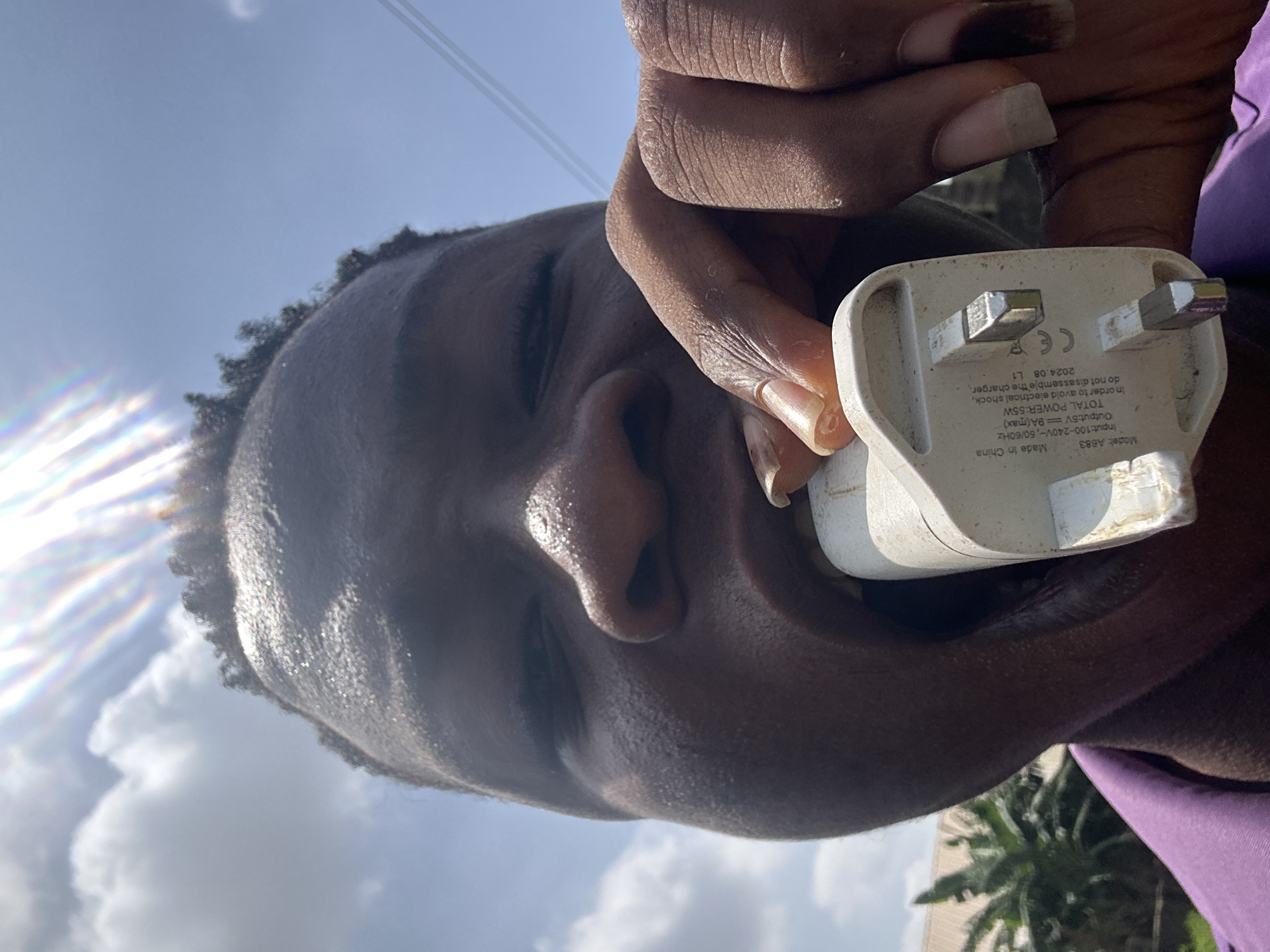 The image is a low-angle selfie taken outdoors in Ikorodu, Nigeria, on a bright, sunny day. A dark-skinned person is holding a white, three-pronged UK wall plug charger close to the camera, with the sky and a few palm trees visible in the background. The charger's label is partially visible and indicates it is a "Model: A683" made in China, with an input of 100-240V and an output of 5V9A (max). The total power is 55W, and it includes a warning not to disassemble the charger. The date "2024.08 L1" is also printed. The person's face is prominent, slightly sweaty, and turned towards the camera, with the charger positioned near their mouth.