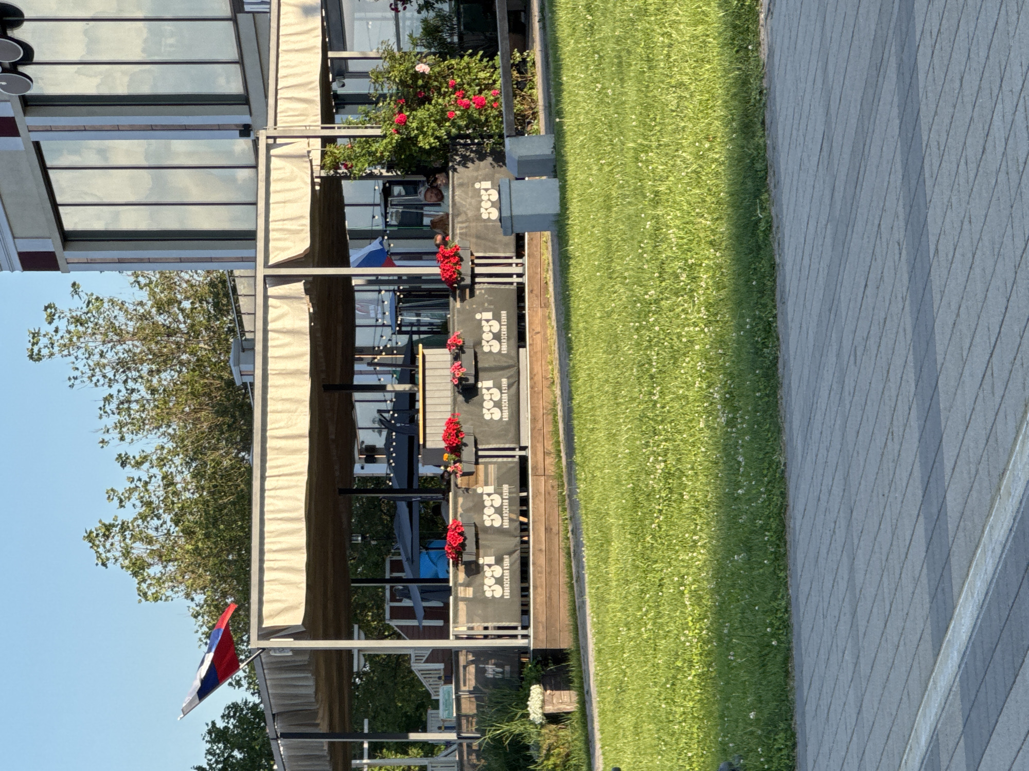 The image shows an outdoor dining area of a restaurant, possibly located in Grushovaya Balka, Russia, based on the presence of the Russian flag. The area is covered by a beige awning supported by metal poles. In front of the awning, a wooden deck is visible, with a railing adorned with banners that display the logo "gogi" and text in Cyrillic. Planter boxes with red flowers are placed along the railing. Beyond the deck, the restaurant building features large, reflective windows. A bush with red roses is prominent on the right side of the image. A patch of vibrant green grass with white clover flowers stretches across the foreground, leading to a paved pathway made of grey tiles. The sky above is clear and blue, with sunlight illuminating the scene.