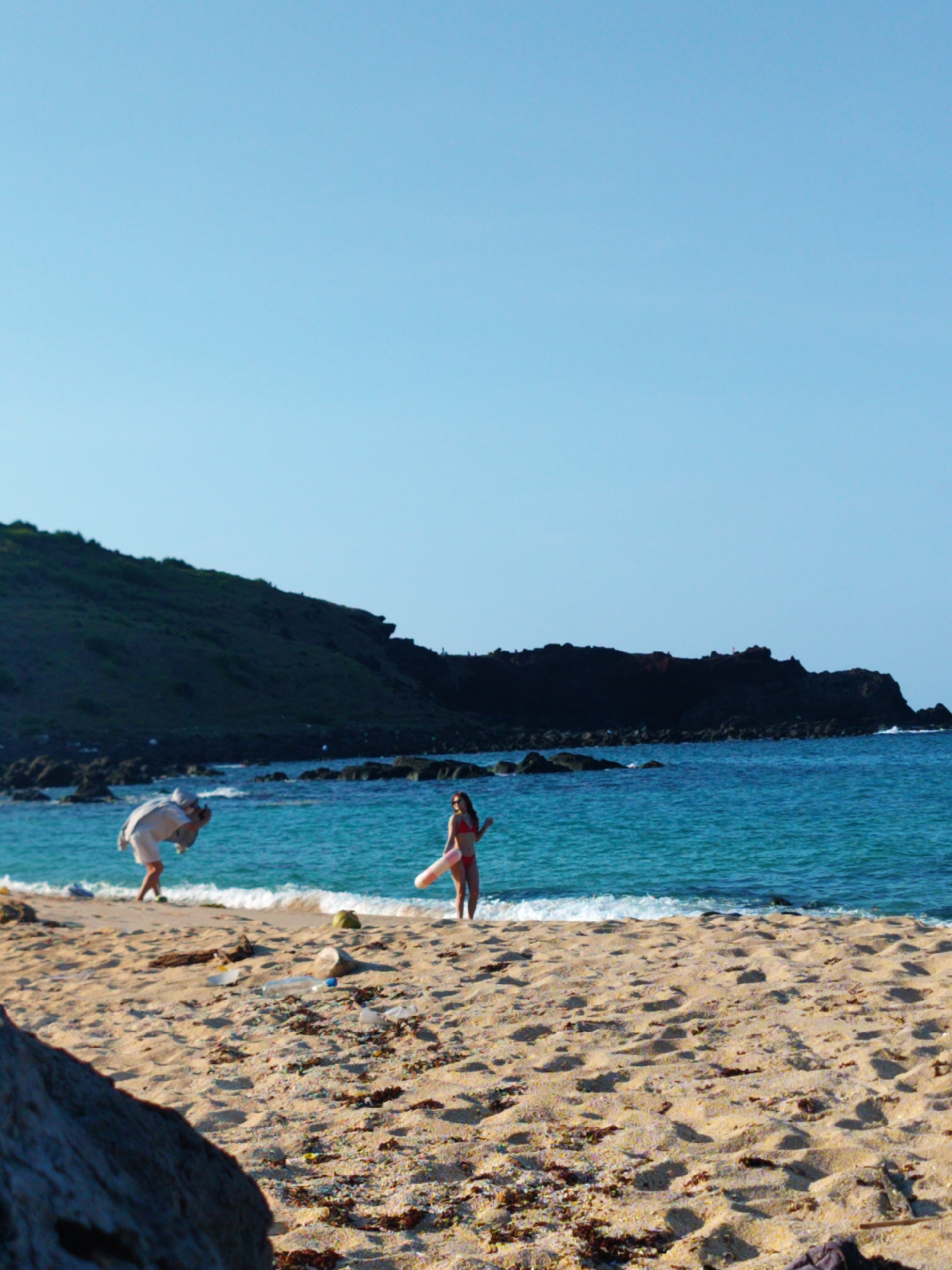 The image shows a beach scene in the Phú Quý Special Administrative Region, Vietnam. In the foreground, there is light brown sand littered with seaweed and debris. A large, grey rock partially obscures the bottom left corner. In the midground, a woman in a red bikini stands near the water's edge, holding a pink and white inflatable tube. To the left, a person wearing light-colored clothing is bent over, possibly taking a picture with a camera or phone. The ocean is turquoise with small waves. In the background, a dark, rocky cliff borders a green hill under a clear, blue sky.