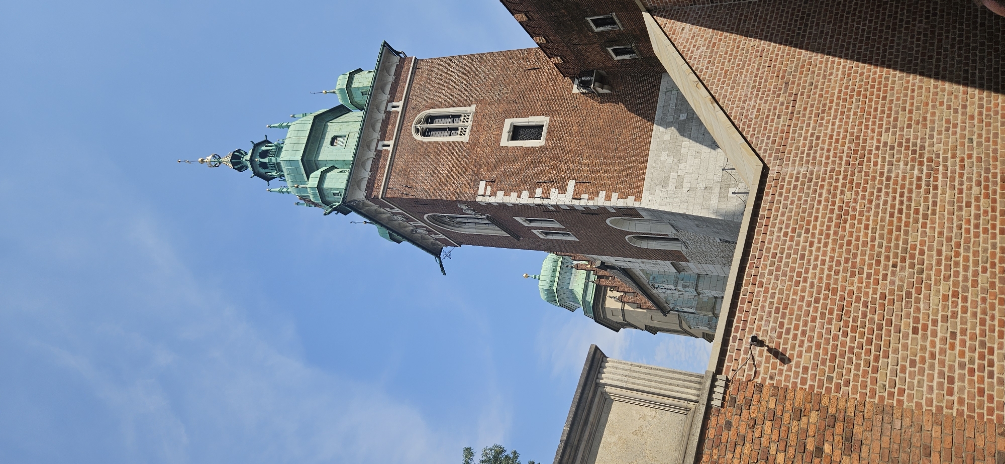 The image shows a close-up view of the Wawel Castle in Krakow, Poland. The scene is dominated by a tall, brick tower with a green, multi-tiered roof. The tower has several windows, some of which are arched. The brickwork of the tower is reddish-brown, and it features white stone accents around the windows and corners.
In the foreground, there is a low brick wall, also made of reddish-brown bricks. This wall casts a shadow, indicating the direction of the sun. Beyond the main tower, another part of the Wawel Castle complex is visible, featuring a green dome. The sky is clear blue with a few wispy clouds. The perspective suggests the photo was taken from within the castle grounds, looking upwards at the architecture. There are no people visible in the image.