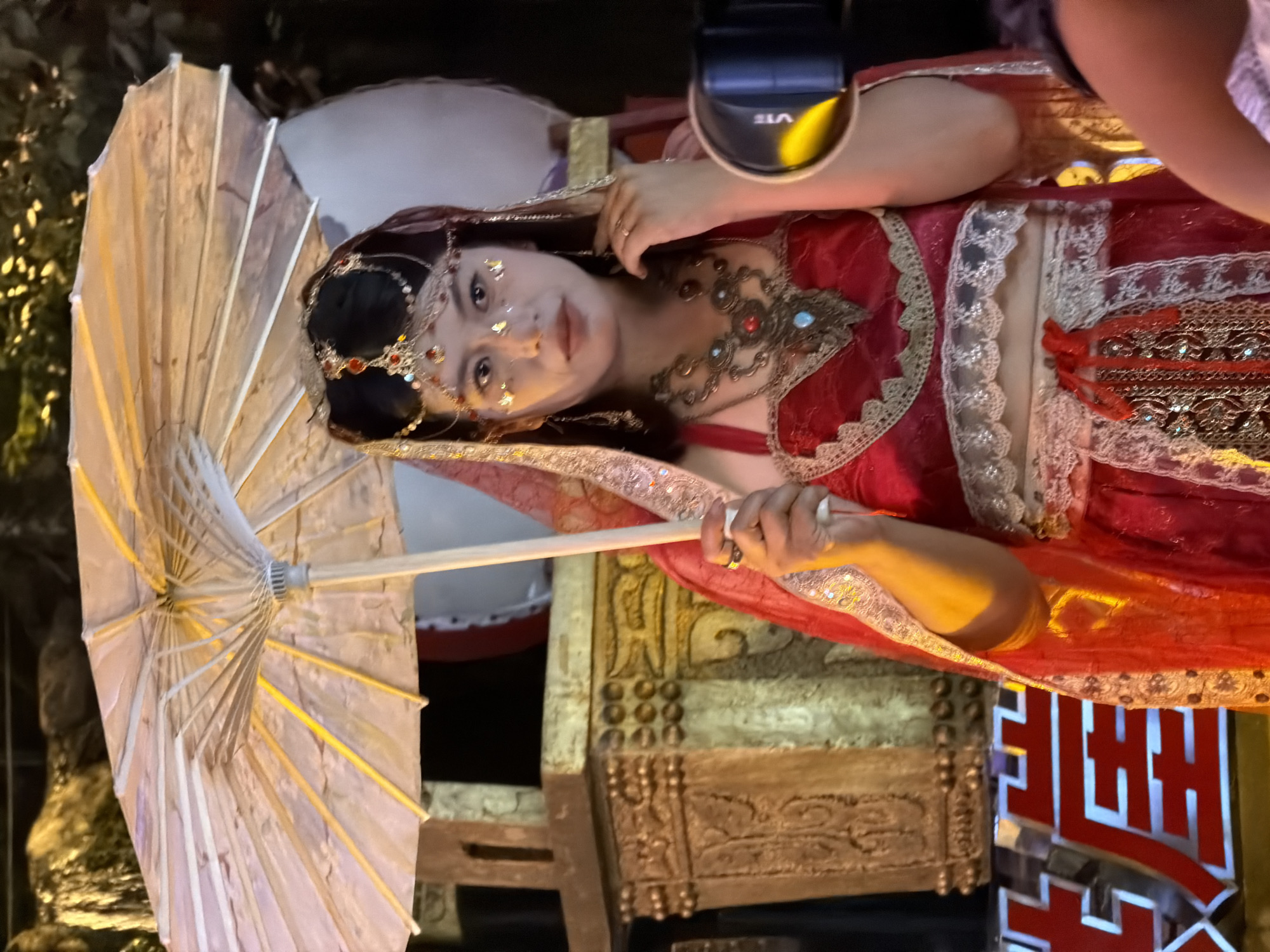 In Yuzhong District, China, a person dressed in a traditional red and gold outfit, adorned with elaborate jewelry, holds a white paper umbrella. The person has intricate makeup and a jeweled headdress. The background features a decorative golden structure with carvings and what appear to be large drums. The lighting suggests an evening or indoor setting, possibly during a cultural event or performance.