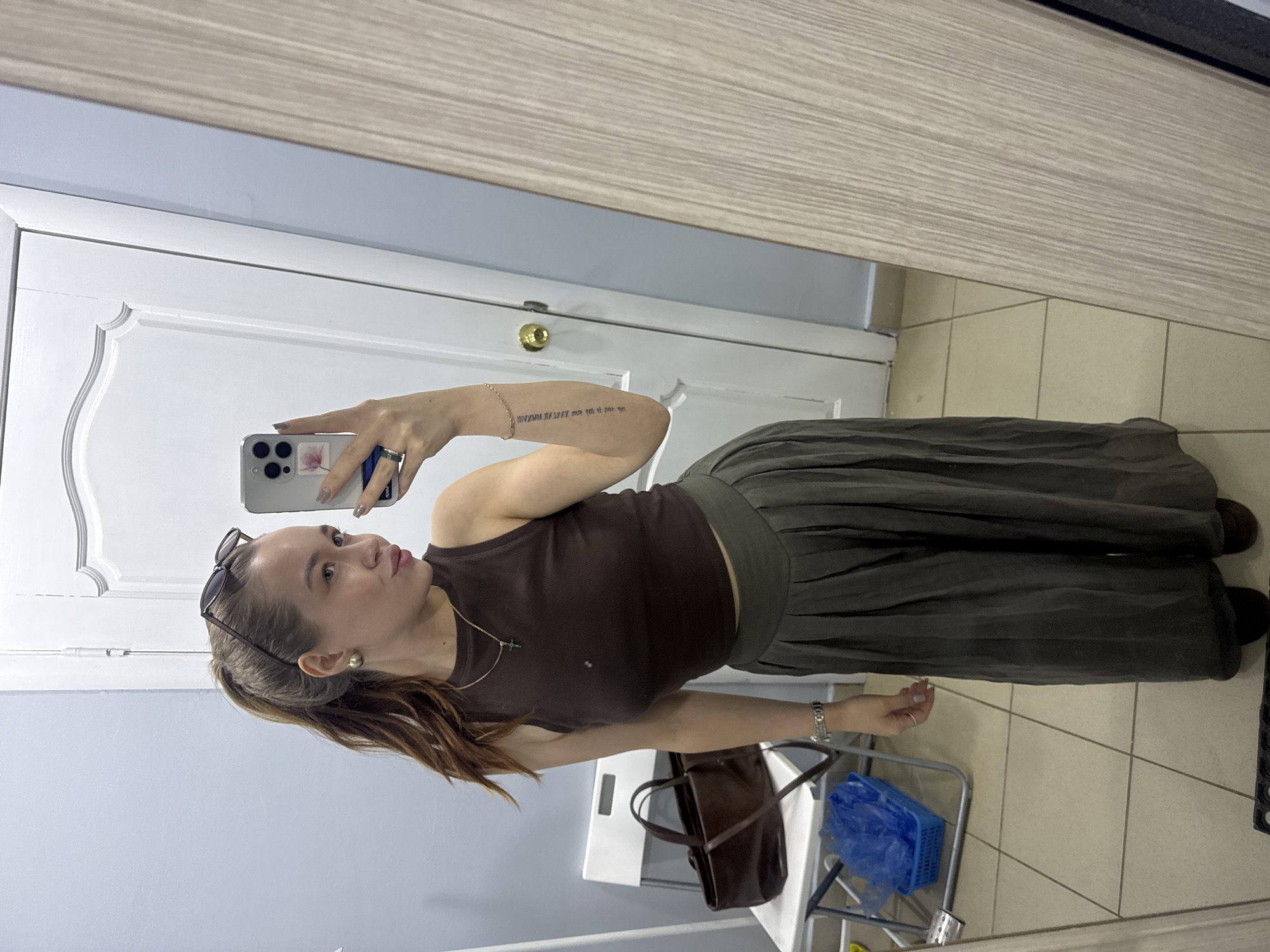 A woman is taking a selfie in a mirror. She is wearing a brown tank top, olive green palazzo pants, and a necklace with a small cross. Her hair is pulled back with sunglasses, and she has a ring on her finger. She is holding a phone with a floral design on the back.
Behind her is a white door with a gold doorknob, and to her right is a light-colored wall. On the floor, there is a tile pattern, a brown leather bag hanging from a chair, and a blue plastic basket on a small rolling cart.
There is no verifiable information in the image that indicates the location is Chaikovsky, Russia.