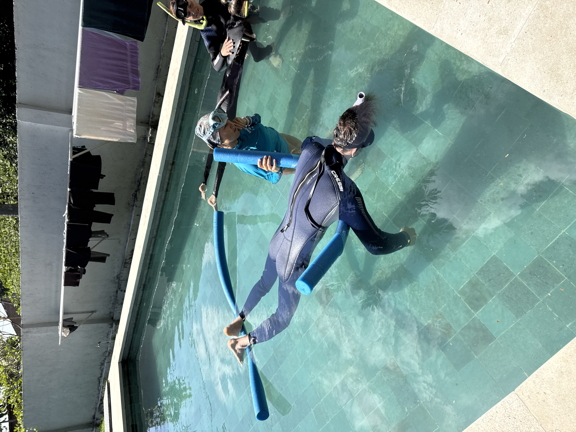 In Nusa Penida, Indonesia, a group of three individuals are engaged in a snorkeling or freediving training session in a swimming pool. One person, wearing a dark blue wetsuit and mask, is floating face down in the water, supported by two blue pool noodles placed under their arms. Their legs are visible at the surface. Another individual, in a lighter blue long-sleeved shirt and a headscarf with goggles, stands in the water holding one of the pool noodles. They are looking down at the floating person. A third individual, in a black wetsuit and mask, is partially submerged in the background, looking towards the other two. The swimming pool has a light-colored tiled bottom. In the background, a white wall with a clothesline stretches across it, with several items of clothing hanging to dry. Trees and a building roof are visible above the wall. The scene is outdoors, under a bright sky.