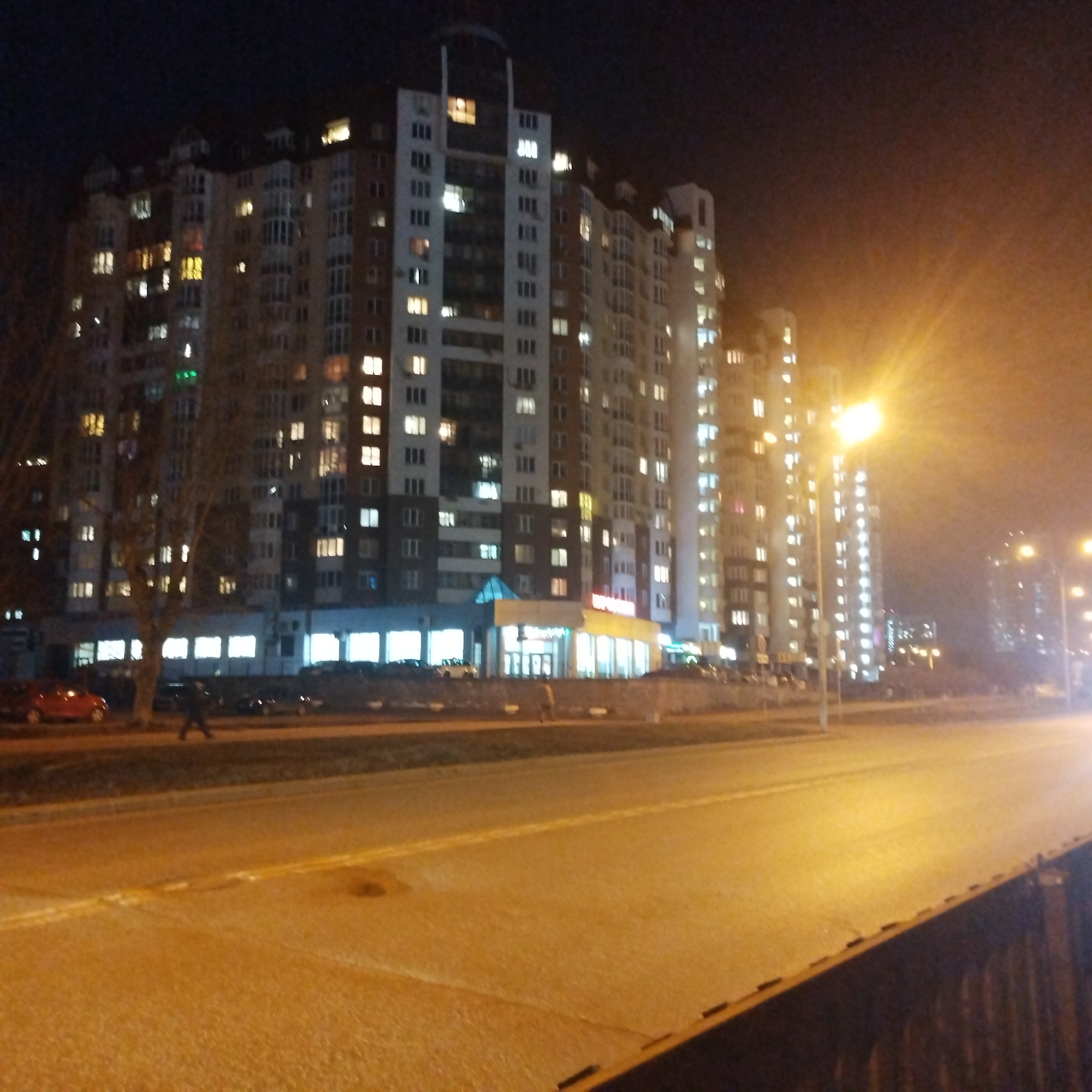 The image shows a night scene of a street in Yekaterinburg, Russia. The street is illuminated by streetlights, which cast a yellow glow. On the left side of the image, there is a large, multi-story apartment building with many windows, most of which are lit up. Cars are parked in front of the building, and a few people can be seen walking on the sidewalk. The street is mostly empty, with only a few cars visible in the distance. The overall atmosphere is calm and quiet.