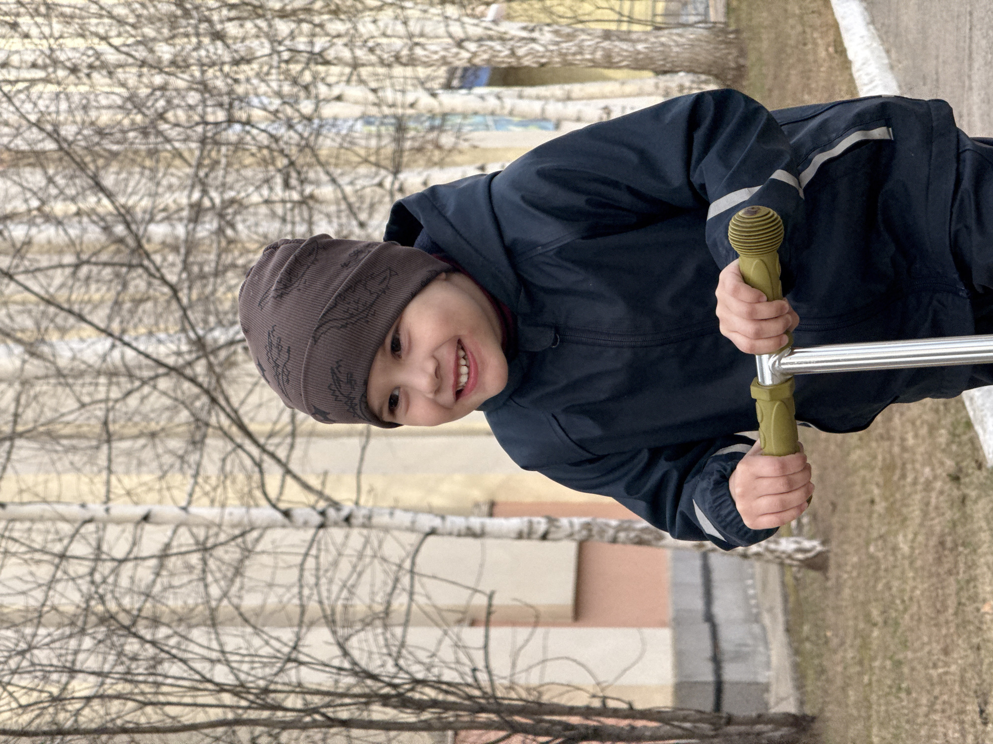 A close-up, slightly tilted portrait captures a young child, likely a boy, smiling broadly. He is wearing a dark blue hooded jacket with light grey reflective stripes on the sleeves and a brown knitted beanie cap featuring a subtle, darker pattern. His face is turned towards the viewer, revealing a wide, joyful smile with visible teeth. The child firmly grips the lime green, ribbed handlebars of what appears to be a scooter or similar ride-on toy, with a shiny metallic bar extending downwards.
The background is dominated by numerous bare deciduous trees, possibly birches, with intricate, leafless branches that suggest late autumn, winter, or early spring. Beyond the skeletal trees, blurred structures with muted tones of beige, light orange, and grey can be seen, likely residential or commercial buildings. The ground appears to be covered with sparse, dry grass and patches of earth. The scene depicts a cheerful outdoor moment, with the bright smile of the child standing out against the cool, muted tones of the environment. The natural light indicates daytime, possibly under an overcast sky. No visible text is present in the image. The setting is consistent with an outdoor area in a location like Chaikovsky, Russia, during a colder season.