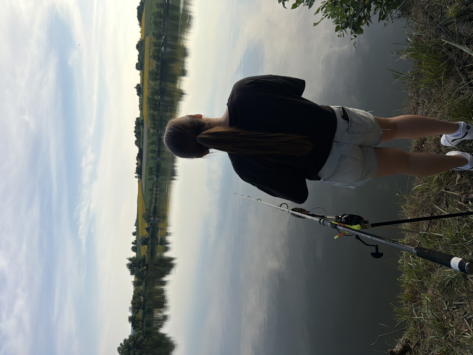 An outdoor, medium shot captures a person from behind, fishing by a tranquil body of water. The individual, with long, dark hair tied in a ponytail, wears a black t-shirt and light-colored denim shorts. A fishing rod is set up on a stand to the left of the person, its tip extending towards the water. The background reveals a calm lake reflecting the sky and a distant shoreline with trees and a field of yellow flowers, characteristic of the rural landscape of Tătărăști, Moldova. The sky above is partly cloudy, with soft daylight illuminating the scene. The foreground consists of grassy, dry terrain with some twigs. The overall impression is one of peaceful solitude and outdoor leisure.