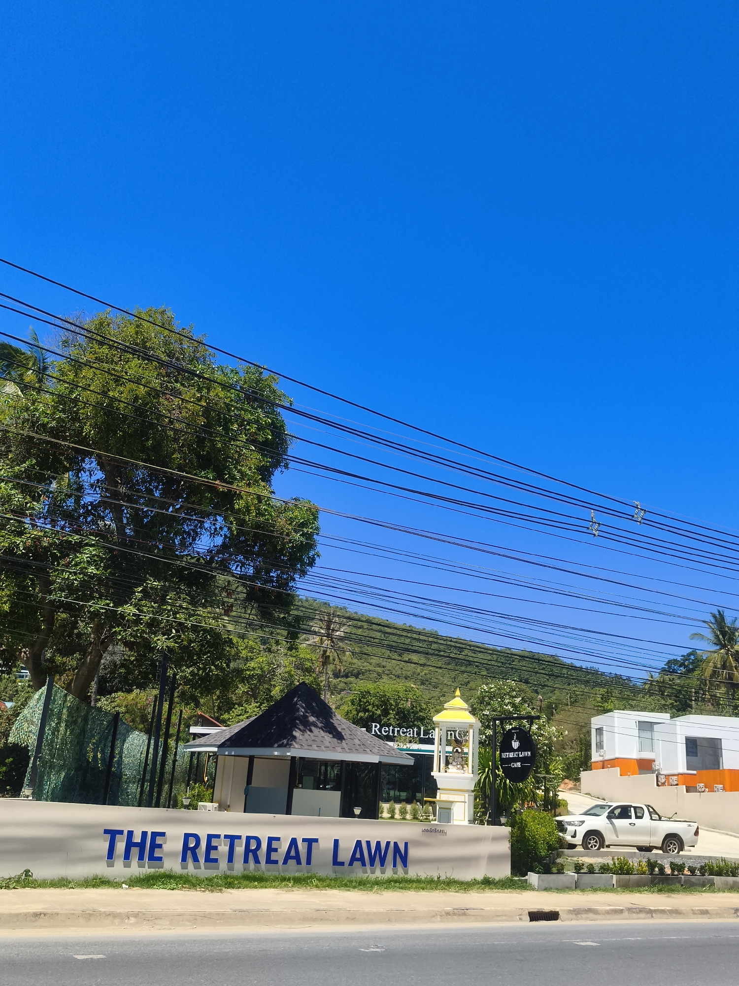 This is a wide-angle, eye-level outdoor shot of a street-level view in Baan Thong Po, Thailand, on a clear, sunny day. The foreground features a light-colored concrete barrier with the blue, three-dimensional letters "THE RETREAT LAWN" prominently displayed. To the right of the barrier, a white pickup truck is parked near a modern building complex with white walls and orange accents.
Behind the barrier, a small, dark-roofed pavilion-like structure stands, with a white wall visible behind it. A short distance to the right of this structure, a yellow tiered monument or decoration is situated, topped with a golden spire. A black circular sign with white text hangs from a pole nearby, reading "RETREAT LAWN CAFE". A large, leafy green tree dominates the left side of the frame, with numerous power lines crisscrossing in front of it and extending across the upper portion of the image. In the background, a lush green hillside rises, dotted with palm trees, and the bright blue sky fills the upper half of the image. The scene appears to be a commercial or hospitality area along a road.