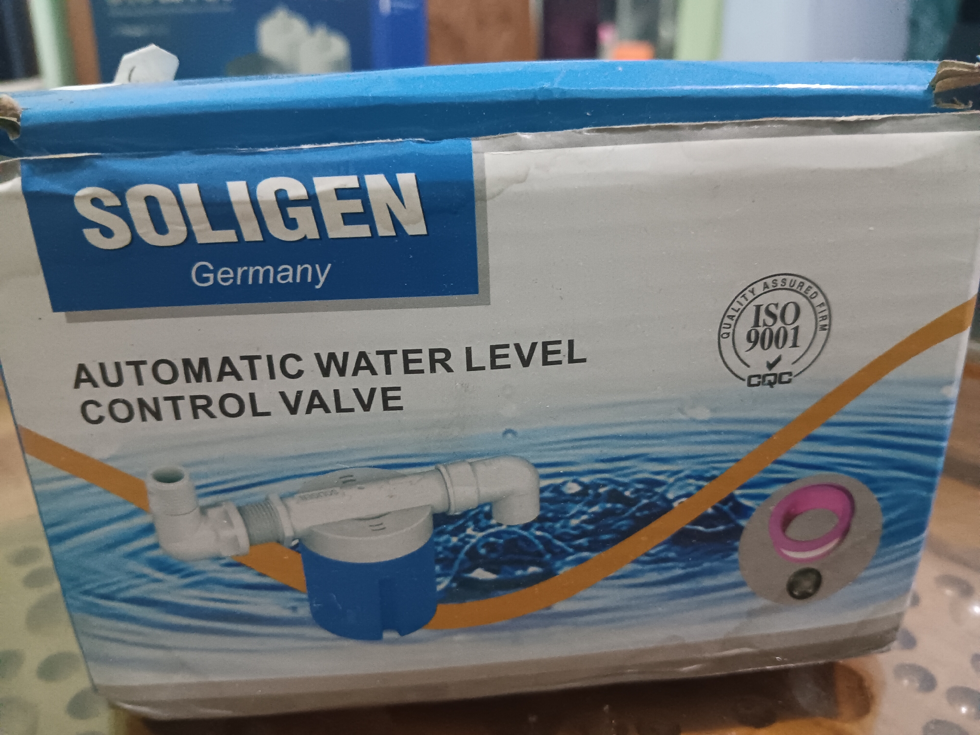 The image shows the box of a Soligen Automatic Water Level Control Valve, made in Germany. The box is predominantly blue and white with orange accents. It features text indicating the brand name "SOLIGEN", "Germany", and "AUTOMATIC WATER LEVEL CONTROL VALVE". A circular "QUALITY ASSURED ISO 9001 CQC" seal is visible on the right side of the box. A diagram of the valve is printed on the lower portion of the box, depicting a blue and white float mechanism connected to a pipe fitting. The box is placed on a patterned surface, possibly a table, in an indoor setting. The background is slightly out of focus but suggests an environment that could be a home or a shop. The image is a close-up of the product packaging.