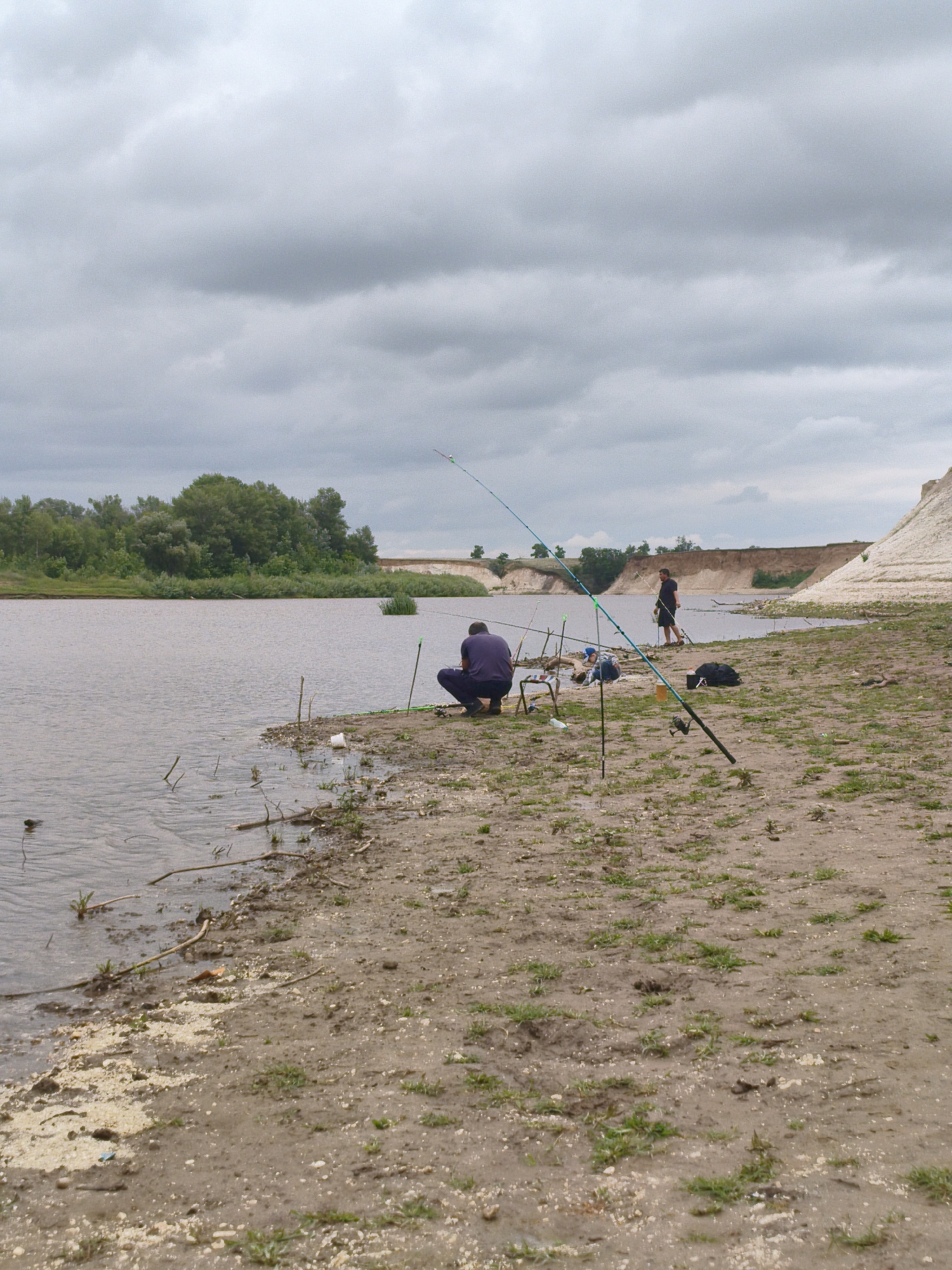In the Ақсуат ауылдық округі of Kazakhstan, on a cloudy day, two individuals are fishing by the river. The scene depicts a sandy and muddy riverbank with sparse green vegetation. In the foreground, a person in a dark purple shirt is squatting, facing the water, with fishing rods and equipment nearby. Further back, another person in a dark t-shirt and shorts stands near the water's edge, also fishing. The river is calm, reflecting the overcast sky. The opposite bank features a sandy cliff face with some greenery at its base and a distant line of trees. Several fishing rods are planted in the ground along the bank, indicating active fishing. A black bag and other fishing accessories are visible on the ground next to the squatting angler.