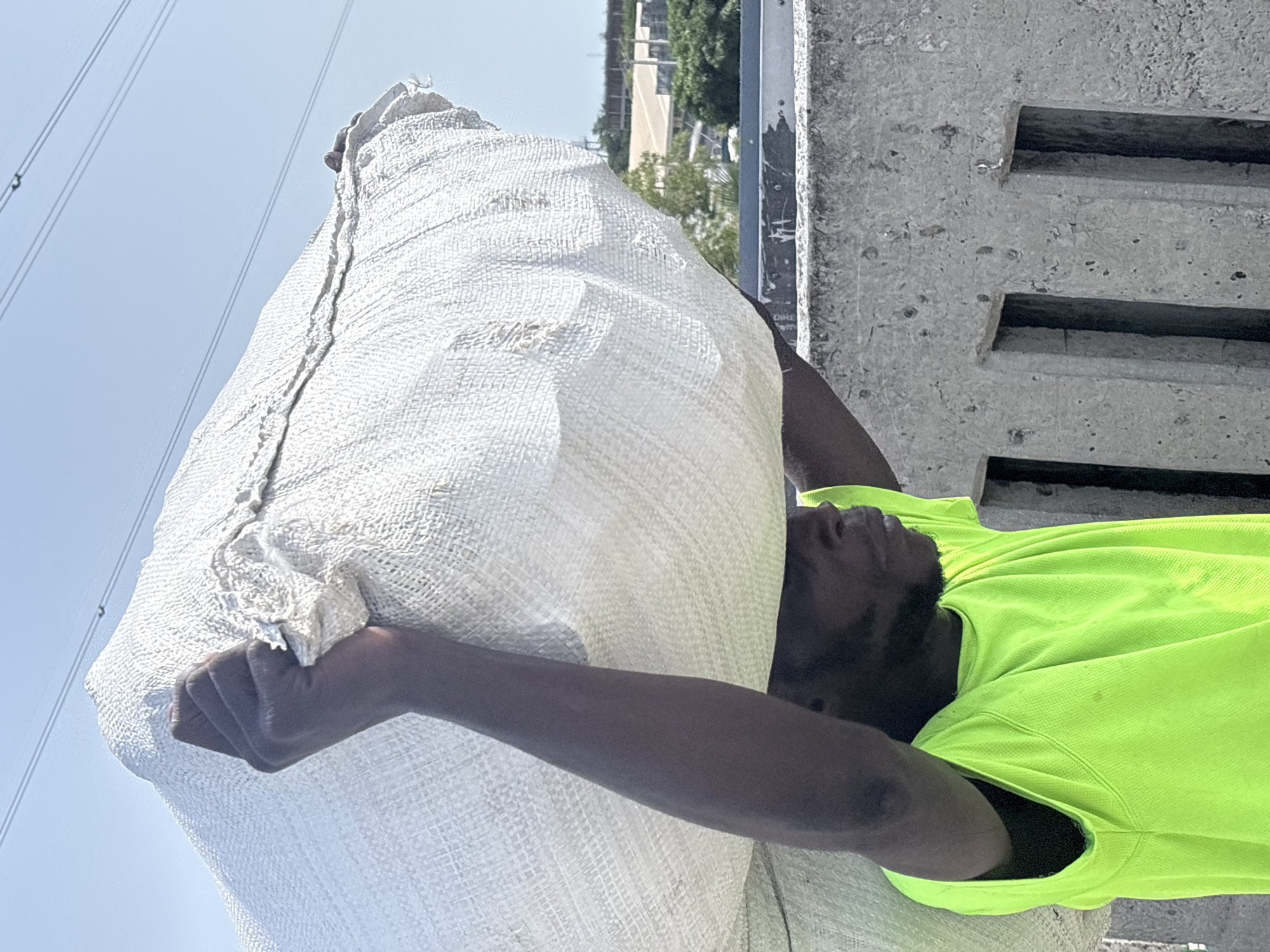 A dark-skinned man in a neon green shirt is carrying a large white sack on his back in Igboefon, Nigeria. He is standing next to a concrete structure with horizontal slots. Power lines are visible in the background.