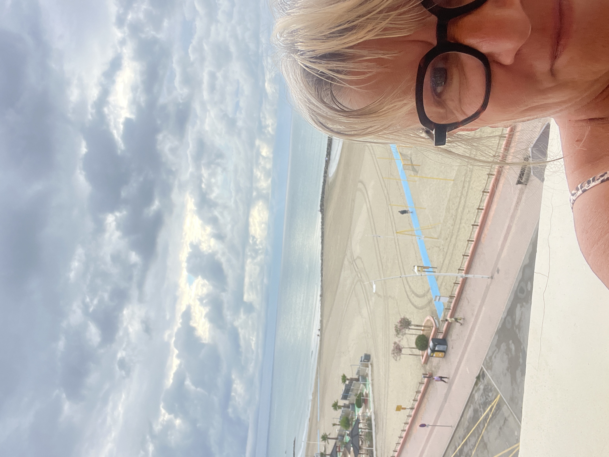 The image shows a woman in the foreground, facing the camera and looking slightly to her right. She has blonde hair and is wearing dark-rimmed glasses and a patterned strap of clothing over her left shoulder. Behind her, a wide, sandy beach stretches towards a calm, blue sea under a cloudy sky. A paved promenade runs along the edge of the beach, with a few people walking or standing. There are some palm trees and shaded structures on the promenade. The overall scene suggests a seaside location in Palavas-les-Flots, France, possibly from a balcony or elevated vantage point.