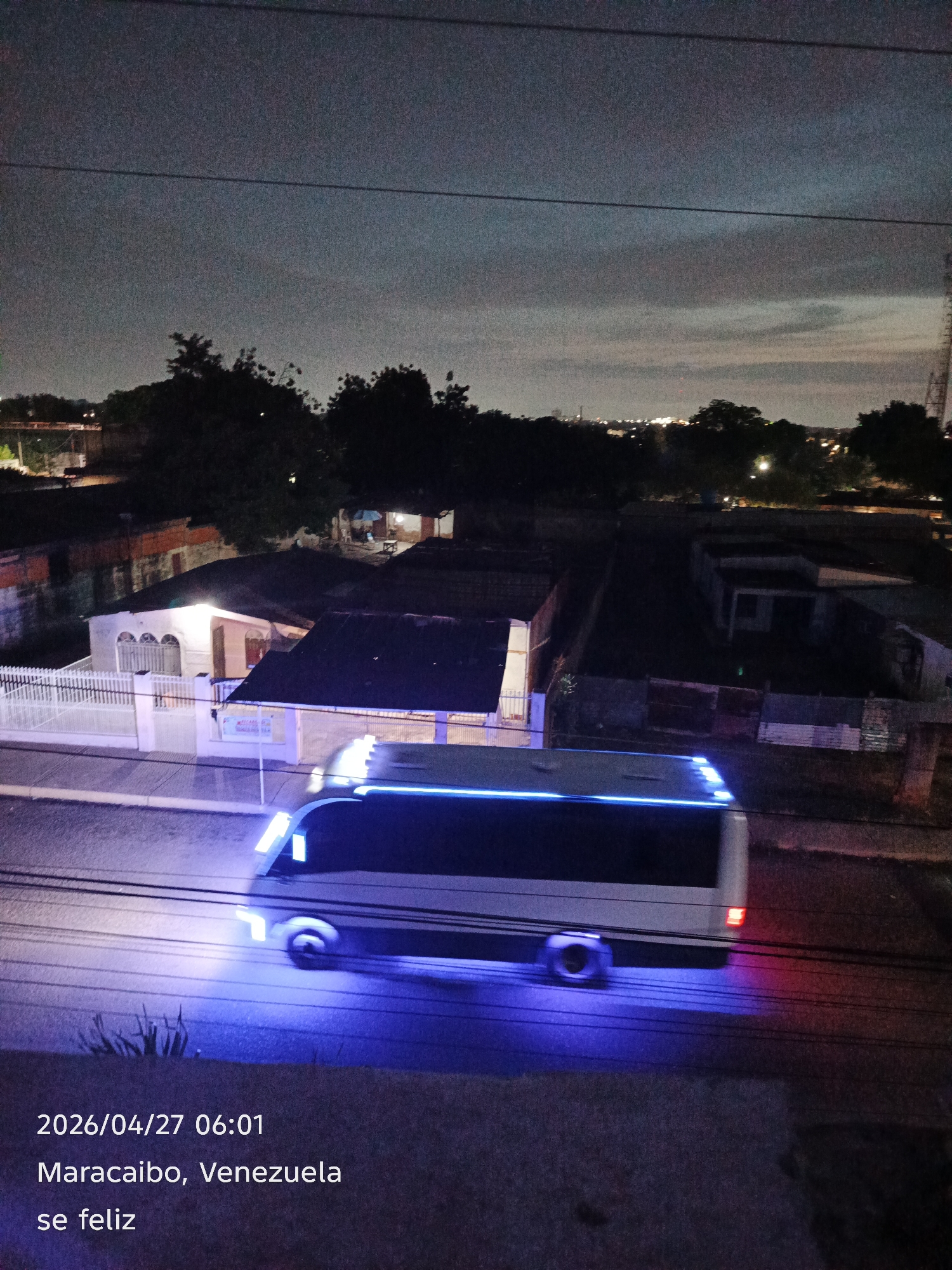 The media file captures an urban street scene at dawn or early morning, specifically at 06:01 on April 27, 2026. The primary subject is a white bus with distinctive blue underglow lights, traveling on a road, evidenced by motion blur. A single red taillight is visible at the rear of the bus.
The scene is set in a residential area, with several houses visible on both sides of the street. Some houses have their interior or porch lights on, casting warm glows. These buildings appear to be modest, with varied architectural styles, some featuring white fences and gated entrances. Lush dark trees are scattered throughout the background and between houses. In the distance, faint lights suggest a broader city landscape under a mostly dark, somewhat cloudy sky. Power lines traverse the upper portion of the frame.
The overall atmosphere suggests the quiet beginning of a day. The artificial blue light of the bus contrasts with the natural dimness of the pre-sunrise sky and the warm domestic lights.
Visible text superimposed on the image indicates:
- "2026/04/27 06:01" (date and time)
- "Maracaibo, Venezuela" (location)
- "se feliz" (Spanish for "be happy")