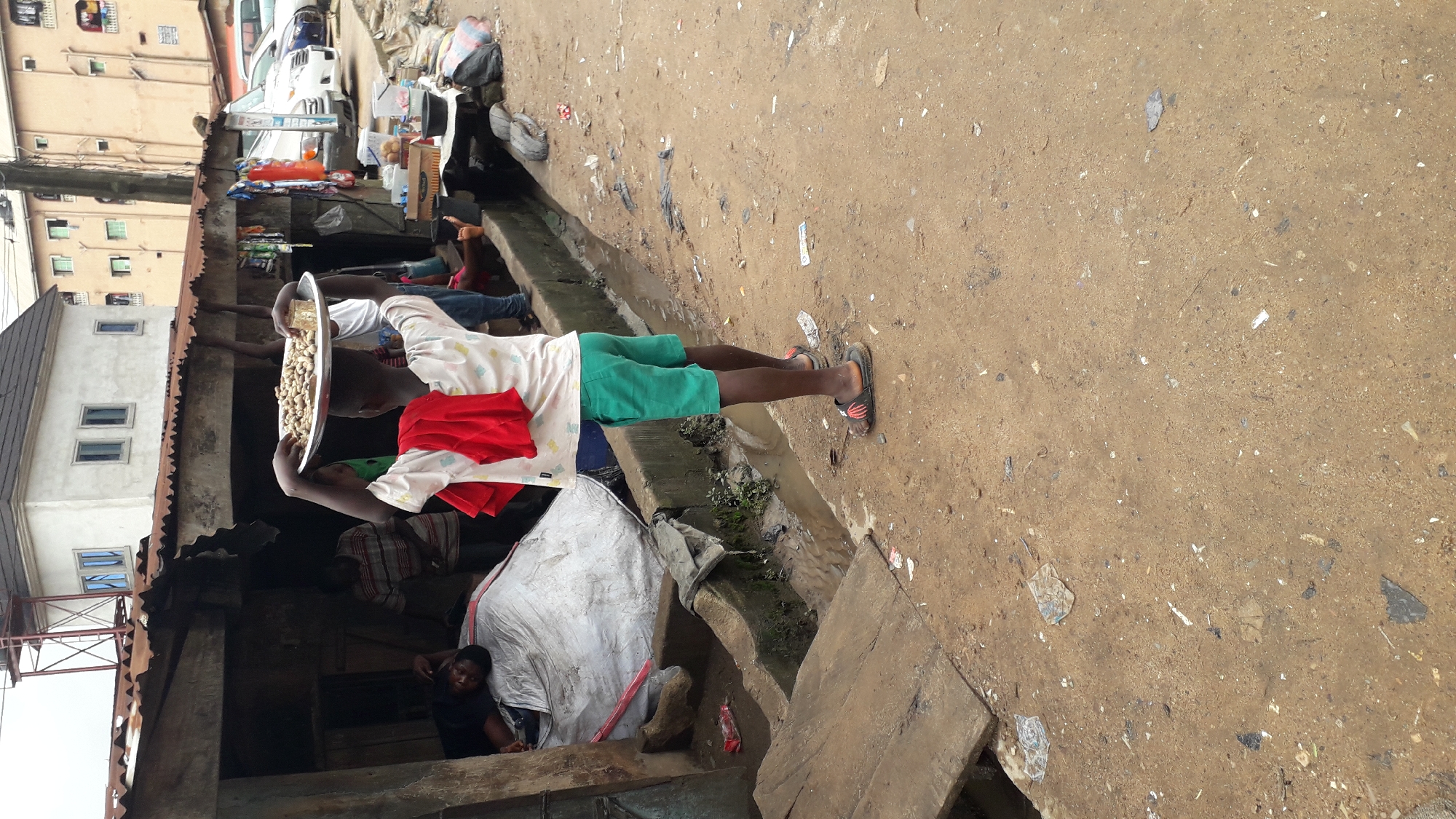 The image depicts a street scene in Onitsha, Nigeria. In the foreground, a young person stands facing away from the camera, carrying a metal tray filled with peanuts on their head. They are wearing a white t-shirt with a colorful pattern, green shorts, and dark sandals. A red cloth is draped over their shoulders. To the left of the young person, a makeshift stall with a corrugated iron roof is visible. Inside the stall, a person is lying down on what appears to be a large, white-wrapped bundle. Other individuals are partially visible behind the stall. The ground is unpaved and dusty, with scattered debris and puddles of water, suggesting recent rain or a poorly drained area. In the background, buildings of varying designs are seen, along with parked vehicles. The overall impression is one of a bustling, outdoor marketplace or residential area.