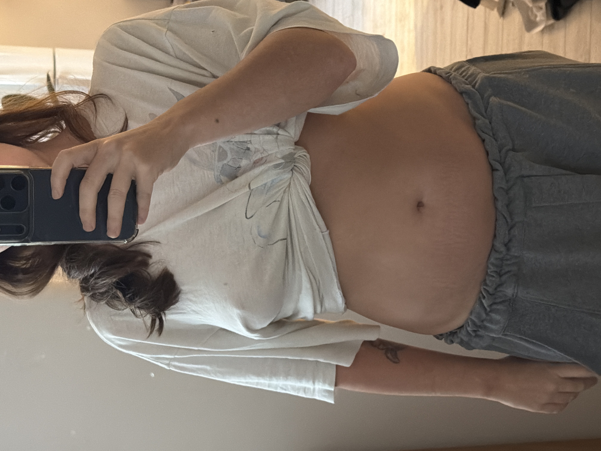 A medium shot depicts a person in an indoor setting, possibly a residence, taking a selfie in a mirror. The person is wearing a light-colored T-shirt tied up to expose their midsection, and gray sweatpants. Their hair is brown. A portion of a smartphone is visible in the person's left hand. Stretch marks are visible on the person's abdomen. A tattoo is visible on the person's right forearm. The background includes a light-colored wall and a wooden floor. Other clothing items are seen partially visible on the floor in the upper right corner of the image. There is no information in the image to determine if the location is in Buenos Aires, Argentina.