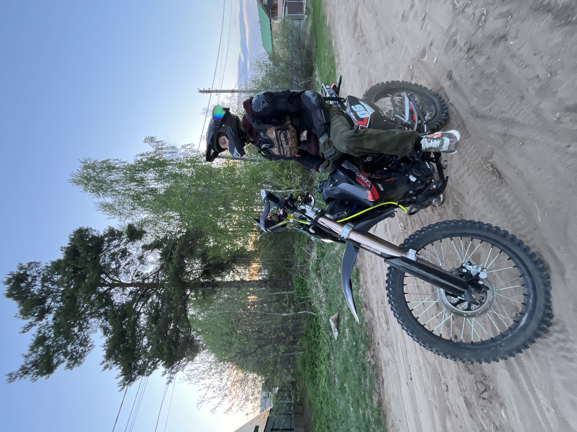 A person wearing a helmet and protective gear is seated on a dirt bike on a dirt road. The bike is black with a number 90 on the side. The person is wearing green pants and multi-colored sneakers. The background shows trees and a building in the distance. The location is likely the city of Chaikovsky, Russia.