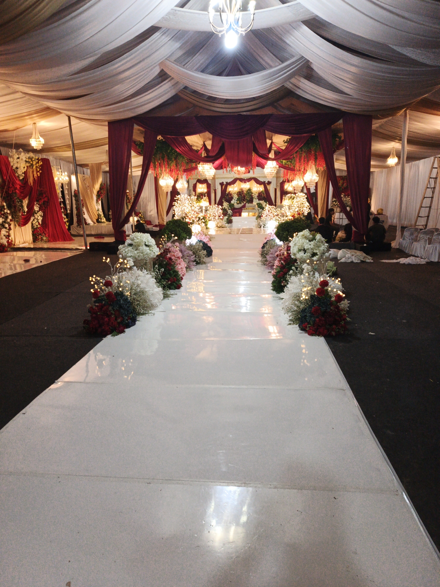This image depicts a decorated event venue, likely for a wedding or formal reception, in Keudealue Ie Puteh, Indonesia. A white, reflective aisle is centrally positioned, leading towards a stage area adorned with red and white drapery, floral arrangements, and chandeliers. The aisle is bordered on one side by dark carpeting and on the other by floral decorations featuring white and red flowers, interspersed with small, illuminated lights. In the background, white tents and draped fabrics create a ceiling and walls for the venue, with additional chandeliers visible. Several people are present in the background, some seated and others near a ladder, suggesting preparation or setup for an event. The overall scene is one of elaborate decoration and anticipation.