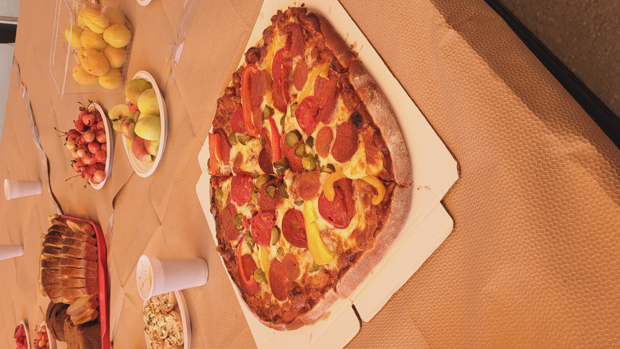 A vertically oriented, high-angle shot shows a spread of food on a textured, tan surface. In the foreground, a large, sliced pizza with pepperoni, tomatoes, peppers, and pickles is the central focus. To the left of the pizza, a container of sliced bread sits in a red basket. Further left, several white paper cups are scattered, one of which is filled with a creamy dip alongside a plate of what appears to be mini quiches or pastries. Behind the bread, a white bowl overflows with bright red cherries. To the right of the cherries, another white bowl contains sliced peaches and apples. In the upper left corner, a clear plastic container holds a pile of yellow-orange apricots. The overall scene suggests an informal gathering or meal. The lighting is bright, indicating daytime, and there are no clear indications of weather or specific location beyond the table setting.