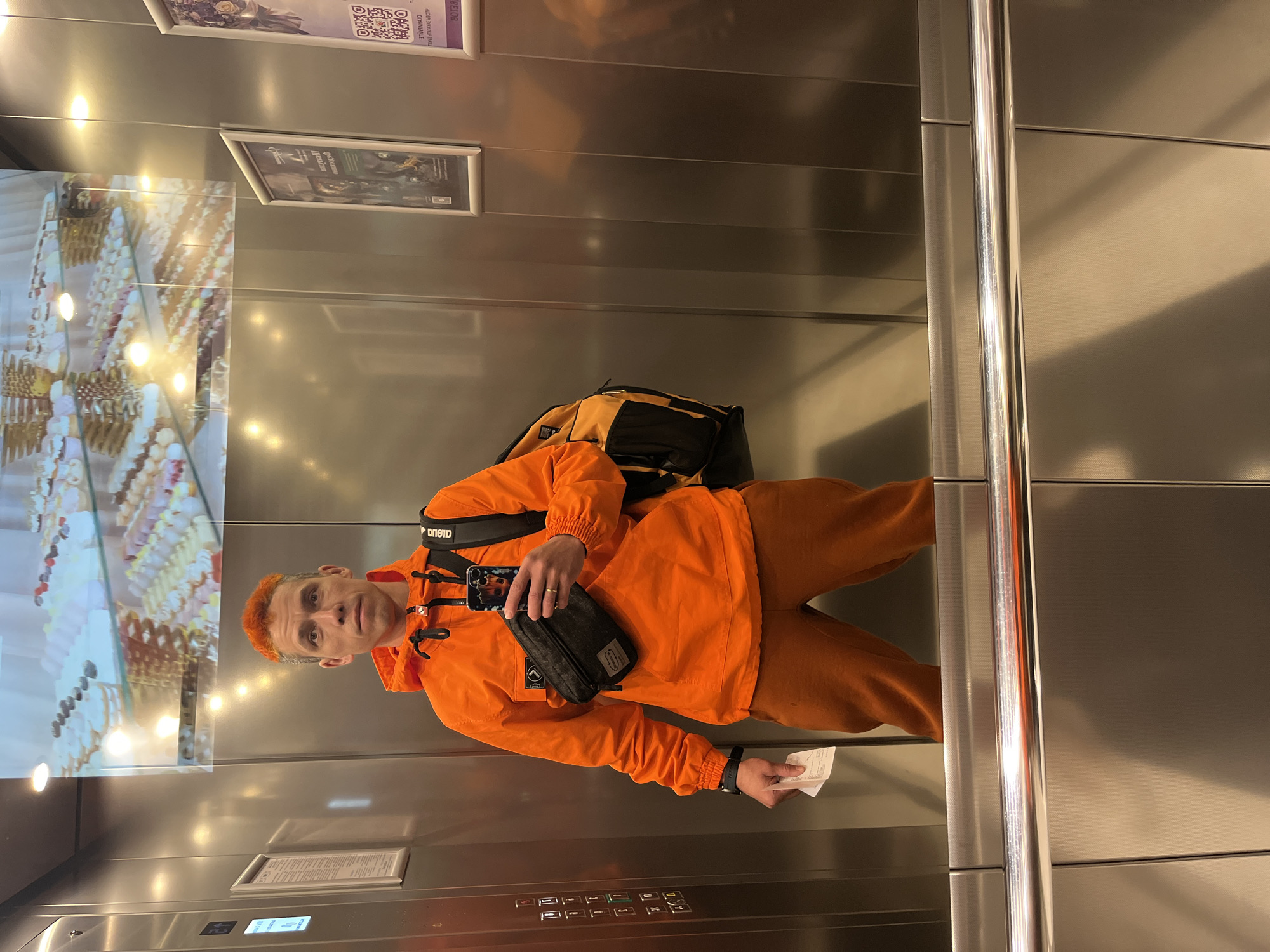 A man stands in an elevator. He is wearing an orange jacket and pants. He has orange hair. He is holding a cell phone in his right hand and a ticket in his left hand. He has a black shoulder bag across his chest. There is a backpack on his back. The elevator has a mirrored wall with a display of pastries. There are also two framed posters on the wall. The elevator has a brushed metal interior. The image was taken in Moscow, Russia.