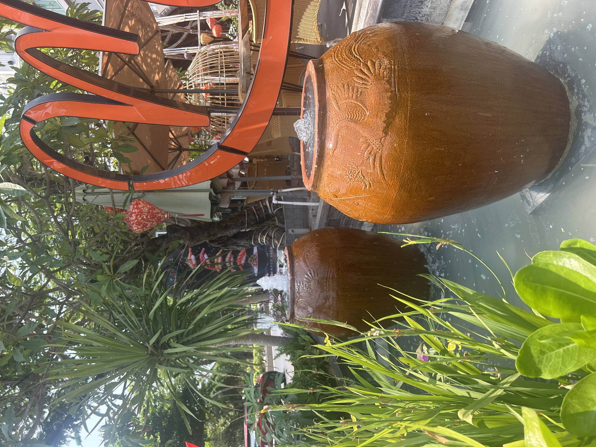 The image depicts an outdoor scene in Nha Trang, Vietnam. The foreground features lush green vegetation, including grassy plants and broader-leafed bushes. A large, textured, brown ceramic jar with water spouting from its top sits in a reflective pool of water. The jar’s reflection is visible in the water. Behind the jar, an orange abstract sign or structure is partially visible, along with a glimpse of outdoor furniture, including wicker chairs and tables with umbrellas. Further back, a tree with hanging decorations is seen. The overall impression is that of a decorative water feature within a landscaped area, likely part of a restaurant or public space.
