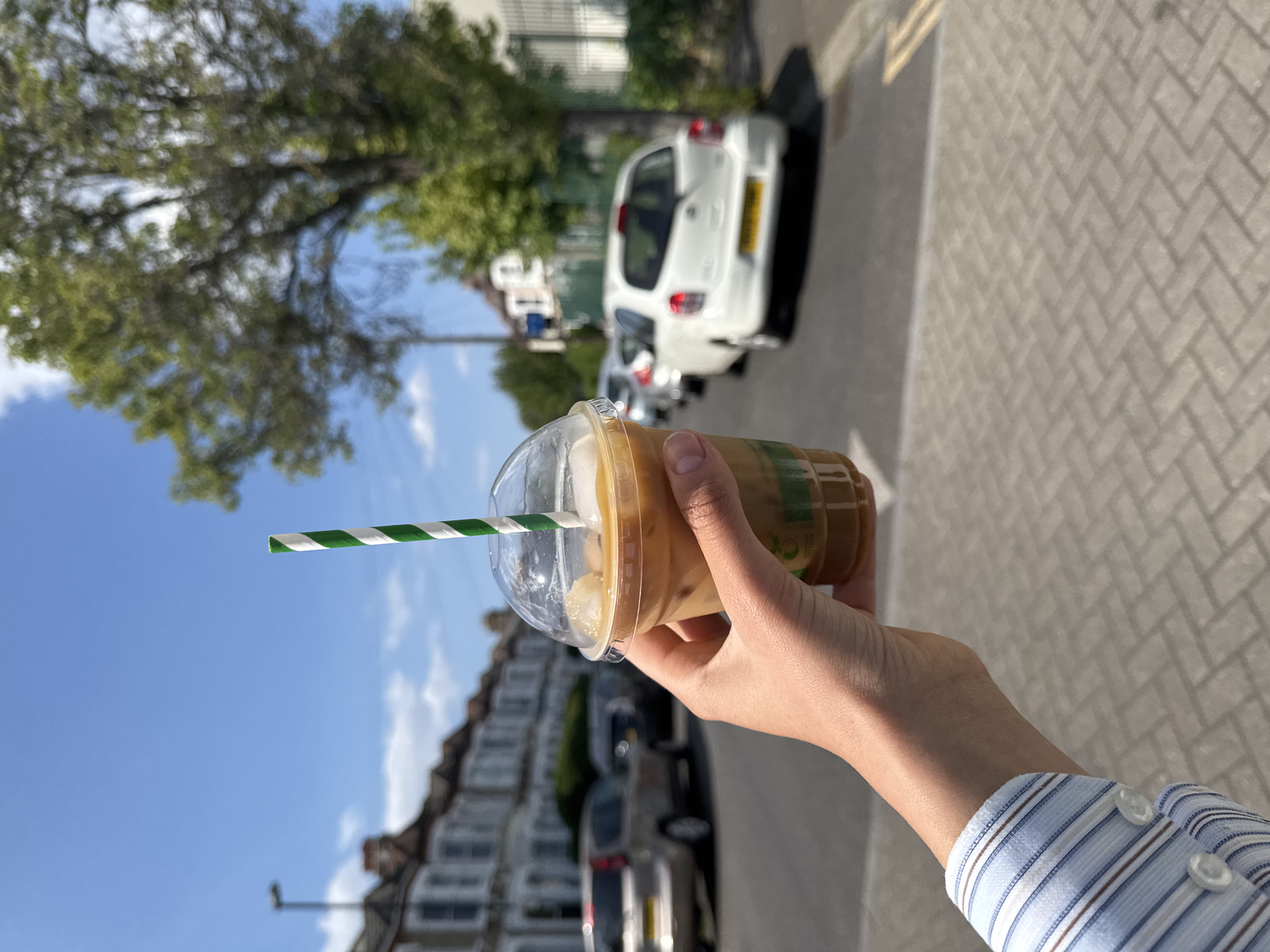 A first-person shot, oriented correctly, captures a person's right hand holding an iced coffee in a clear plastic cup. The cup contains a light brown beverage with ice cubes and a white frothy topping, and a green and white striped paper straw is inserted. The person wears a light blue and white striped shirt with thin brown pinstripes and white buttons on the cuff.
The setting is an outdoor urban or suburban street, likely in London, United Kingdom, given the architecture and visible yellow UK license plate on a parked car. The ground is paved with light-colored interlocking pavers laid in a herringbone pattern. Along the street, several cars are parked, prominently a white hatchback. A row of multi-story residential buildings with traditional architecture lines the street on one side, while lush green trees are visible on the other and in the distance. The sky is bright blue with scattered white clouds, indicating a clear and sunny day, possibly morning or afternoon. No specific activity beyond holding the drink is evident. There is no discernible text on the cup or car registration plate.