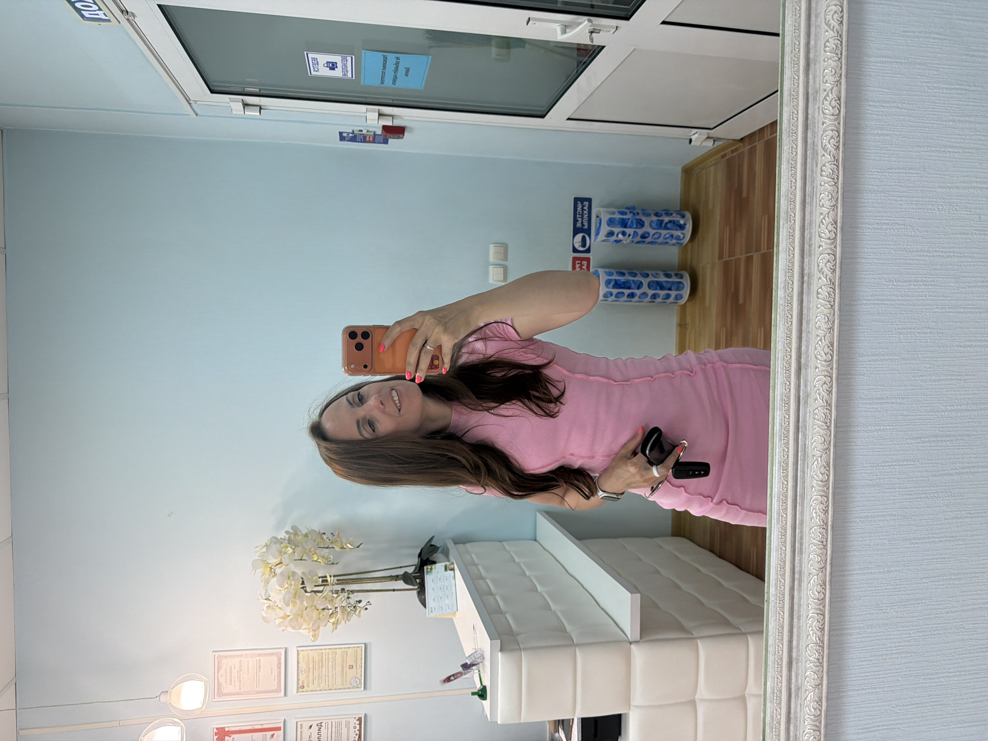 A woman with long brown hair and a pink dress stands in front of a white reception desk, taking a selfie with an orange-cased smartphone. She is holding a black clutch bag and keys in her right hand. The reception desk has a white quilted front and a shelf. Behind the desk, there are several framed certificates or diplomas on the wall. To the right of the reception desk, there is a white door with a glass panel. On the wall near the door, there are blue signs and a red fire alarm. In front of the door, there are two cylindrical white containers with blue plastic bags inside. The floor appears to be made of wood planks and tiles. The overall environment suggests a professional or commercial setting, possibly a salon or office. The location is identified as Birobidzhan, Russia.
