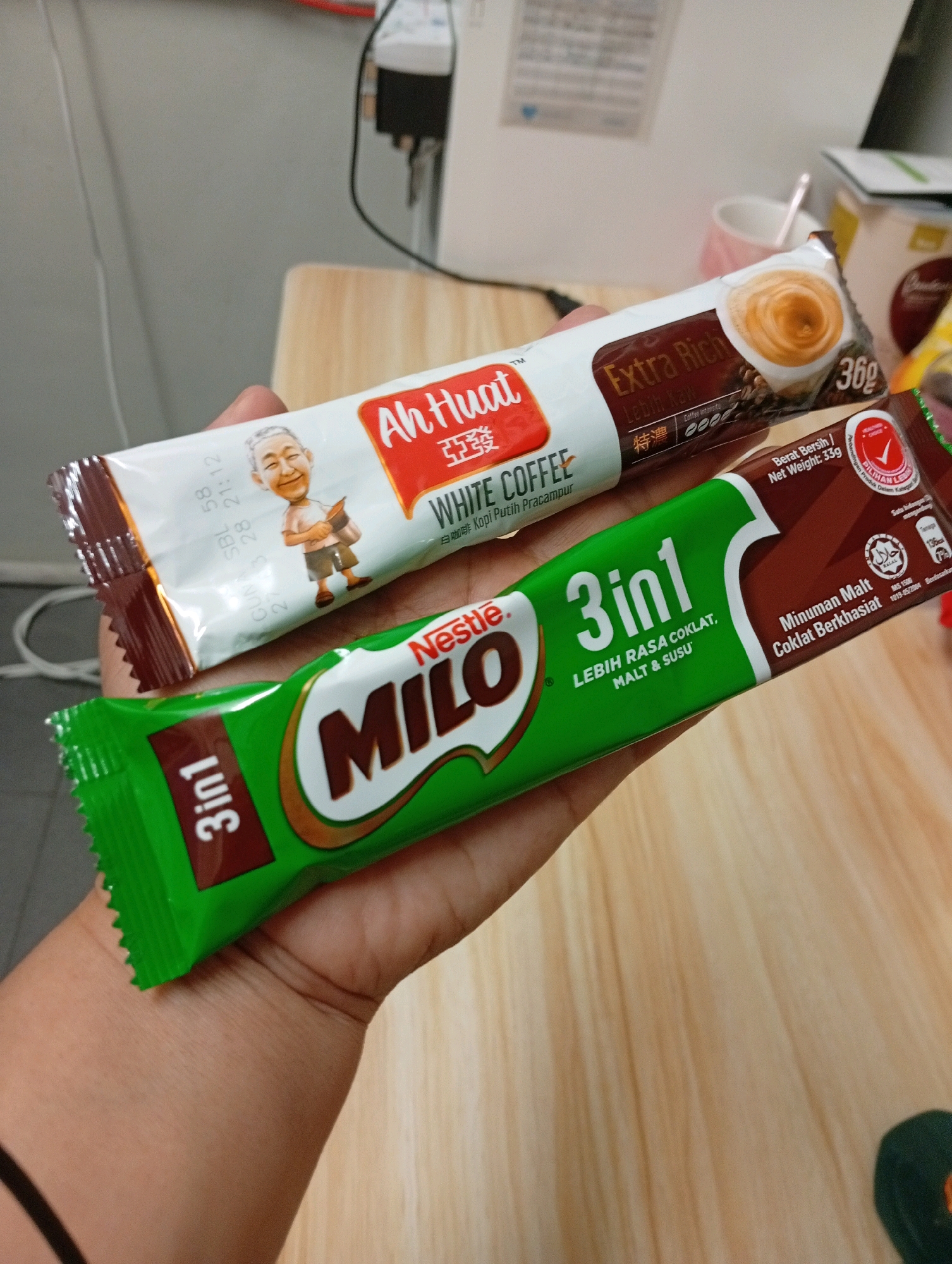 Two packets of instant beverage are held in a person's left hand over a light-colored wooden surface. The upper packet is white with red and brown accents and features an illustration of an elderly Asian man. Text on this packet reads "Ah Huat" and "WHITE COFFEE". The lower packet is bright green and brown and displays the "Milo" logo. Text on the Milo packet reads "Milo 3in1 LEBIH RASA COKLAT, MALT & SUSU". The image appears to be taken indoors, likely in a residential or office setting, possibly within the Selayang Municipal Council area of Malaysia, given the context of the products shown.